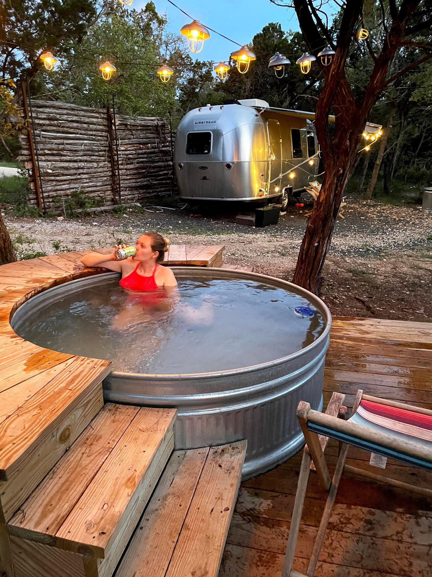 Lydia sits in a hot tub drinking a beer with an airstream in the background. There are hanging lights up above.