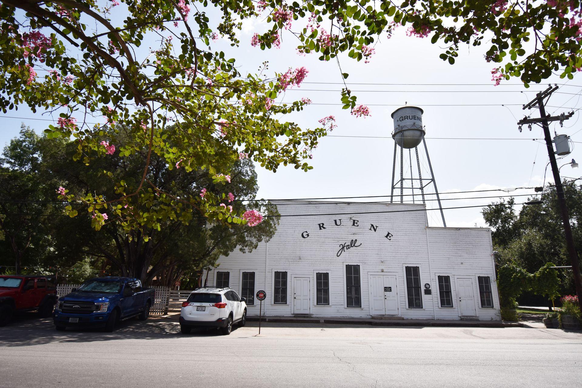 A view of the Gruene Dance Hall from across the street. There are green trees with pink flowers surrounding the building. You can see the Gruene water tower in the background.