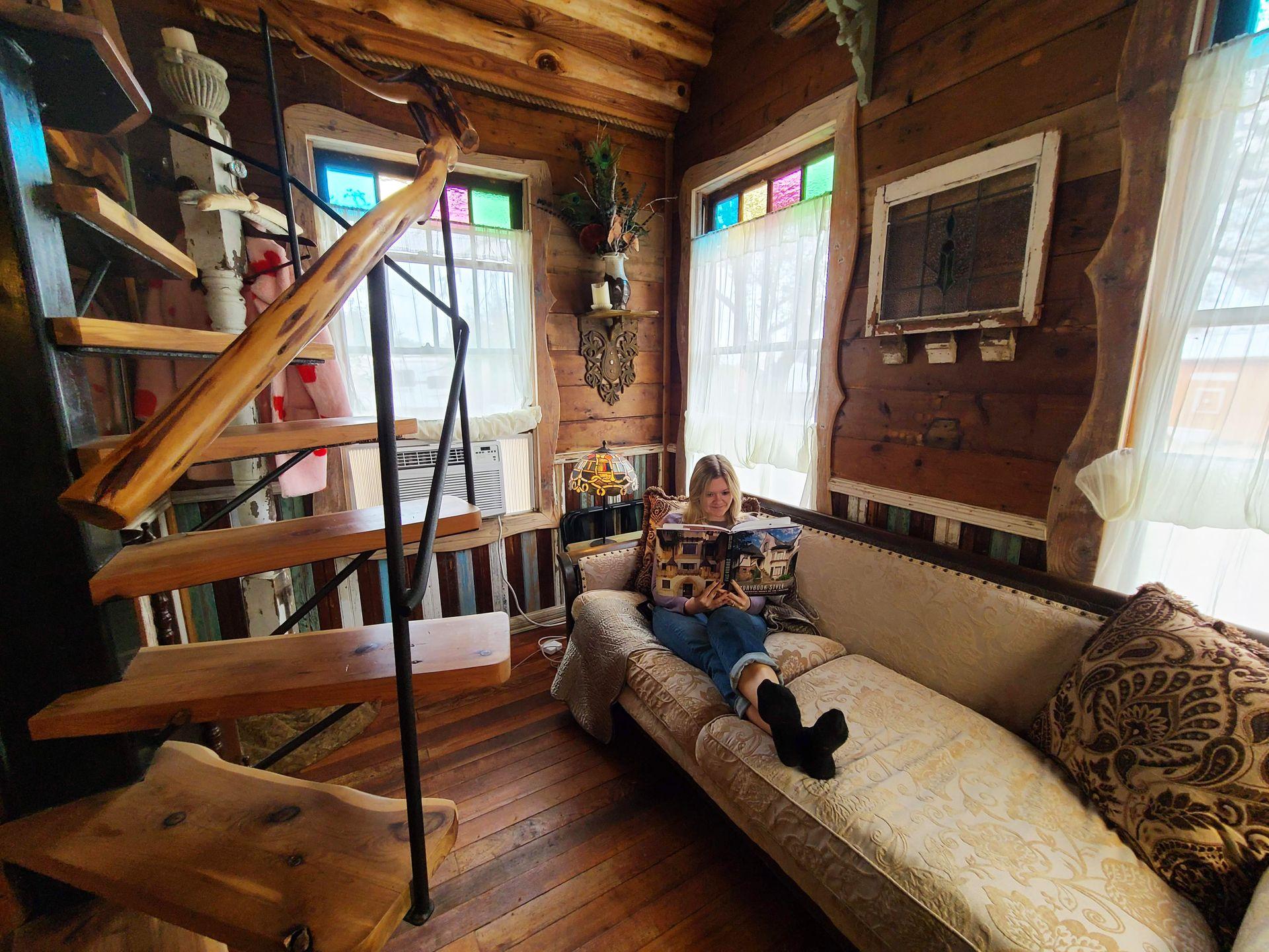Lydia sits on a couch reading a magazine inside of a cottage with stained glass windows, a spiral staircase and vintage furniture.