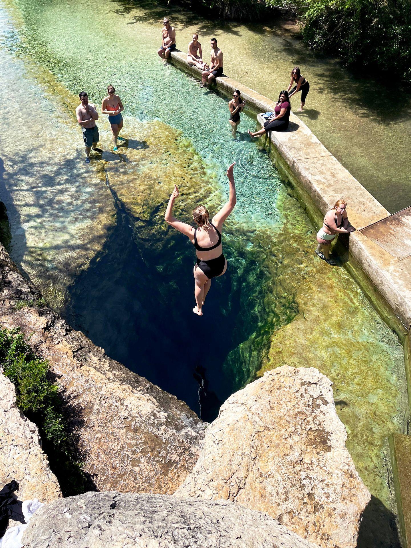 A view from above of Lydia jumping into Jacob's Well. The water is very clear and you can see the underground cave. A few people stand in the shallow water near the well.
