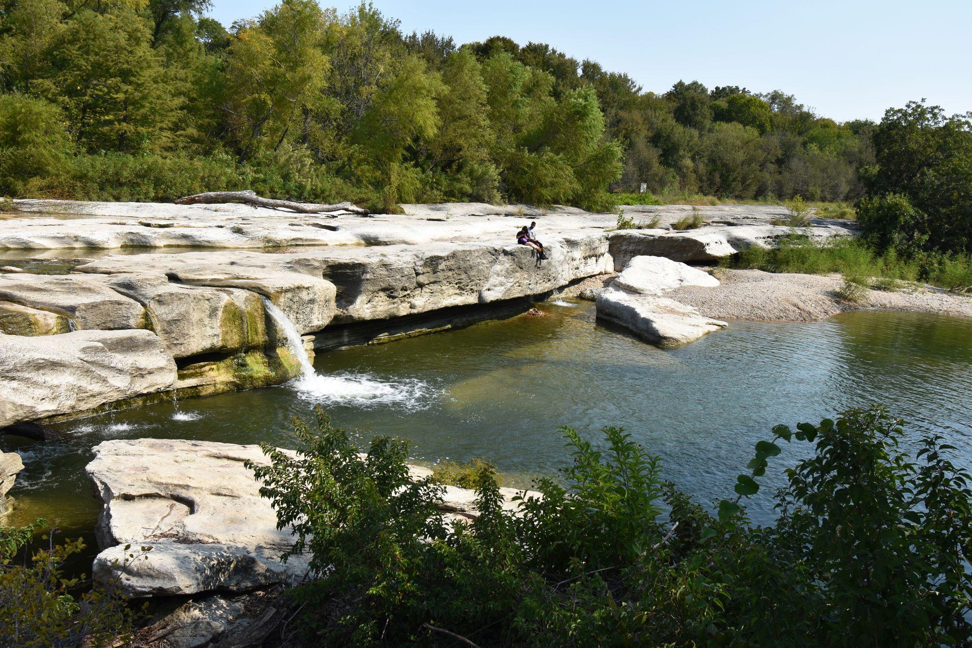 A rock wall with a waterfall at McKinney Falls State Park. The waterfall flows into a swimming area and two people sit on the rocks.