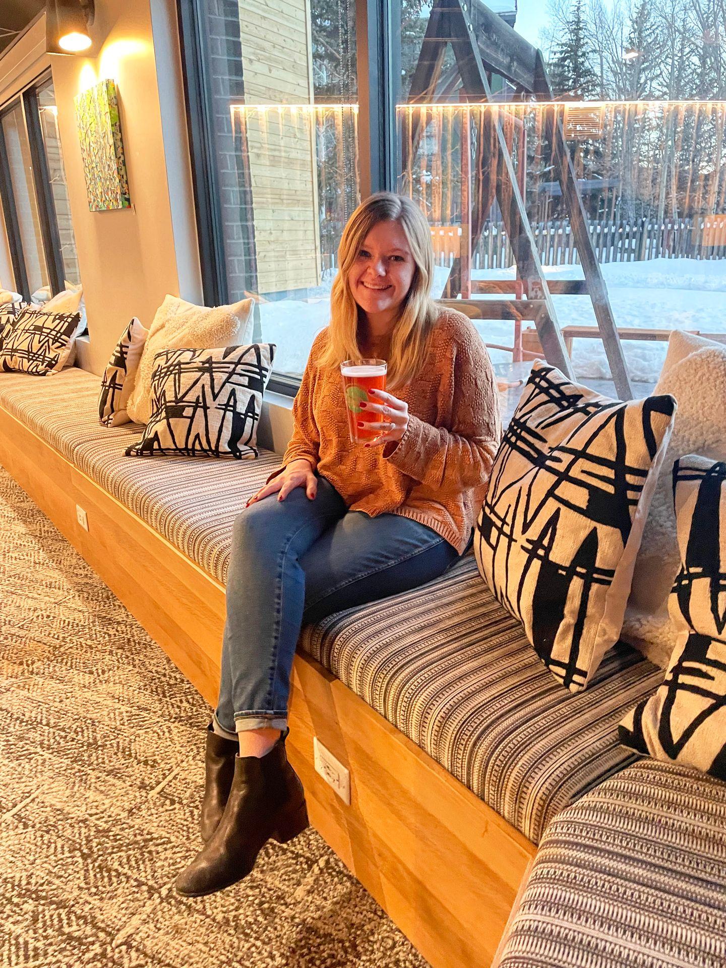 Lydia sitting in the lobby at The Pad holding a beer. There are black and white geometric pillows on the bench surrounding her.