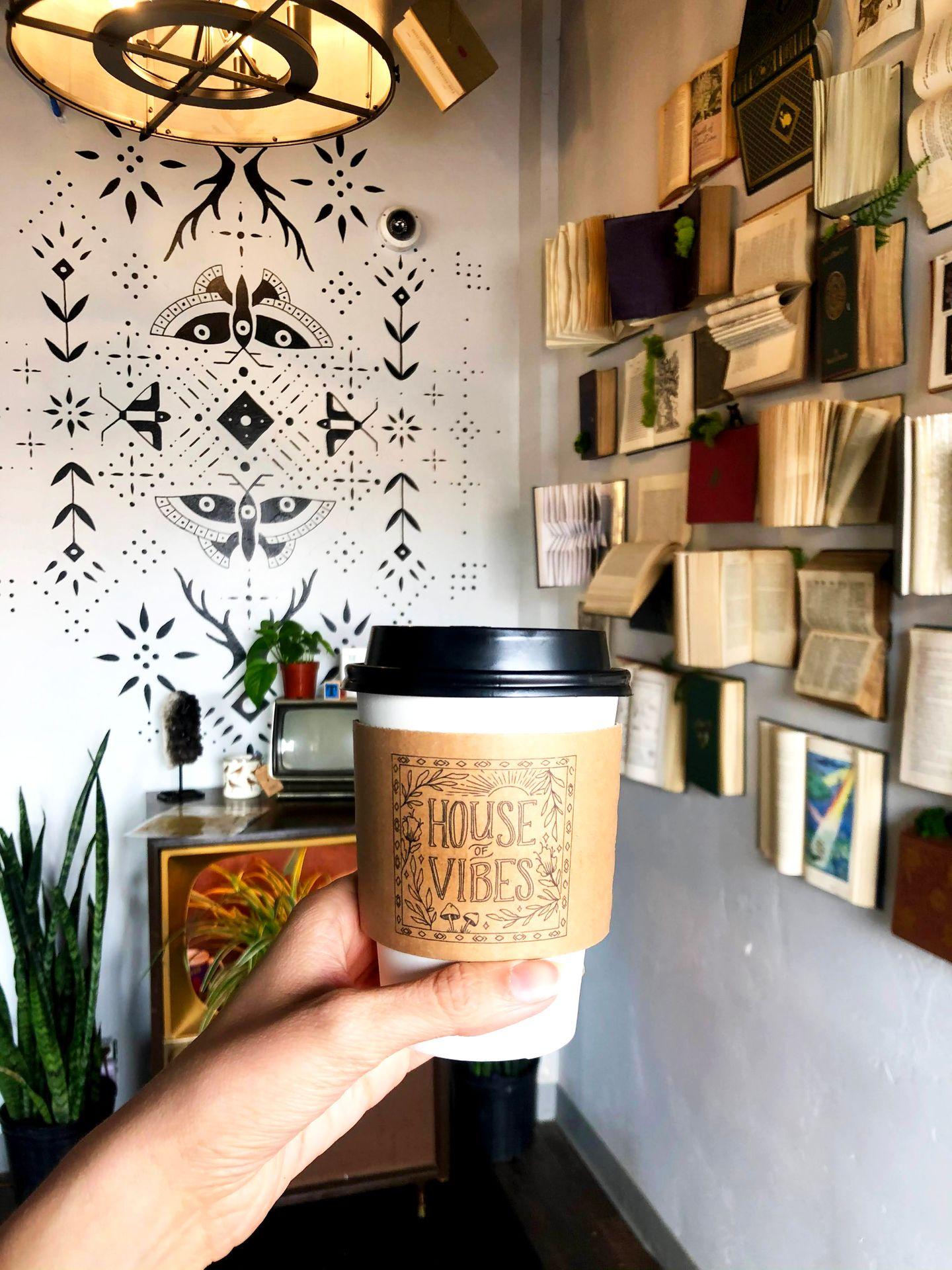 Holding up a coffee in the entry of House of Vibes. The wall is covered in books and a geomtric pattern. There is also a vintage table with some plants.