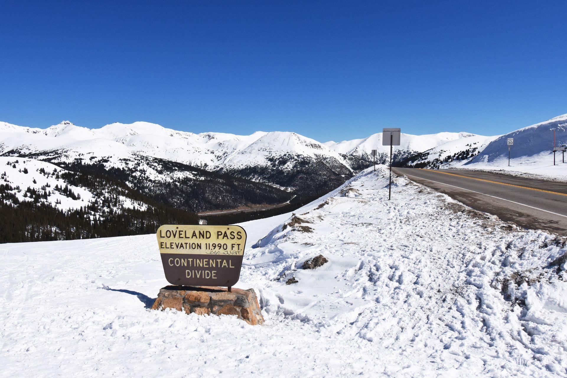 A view of the Loveland Pass sign with snow covered mountains and the road in the background.
