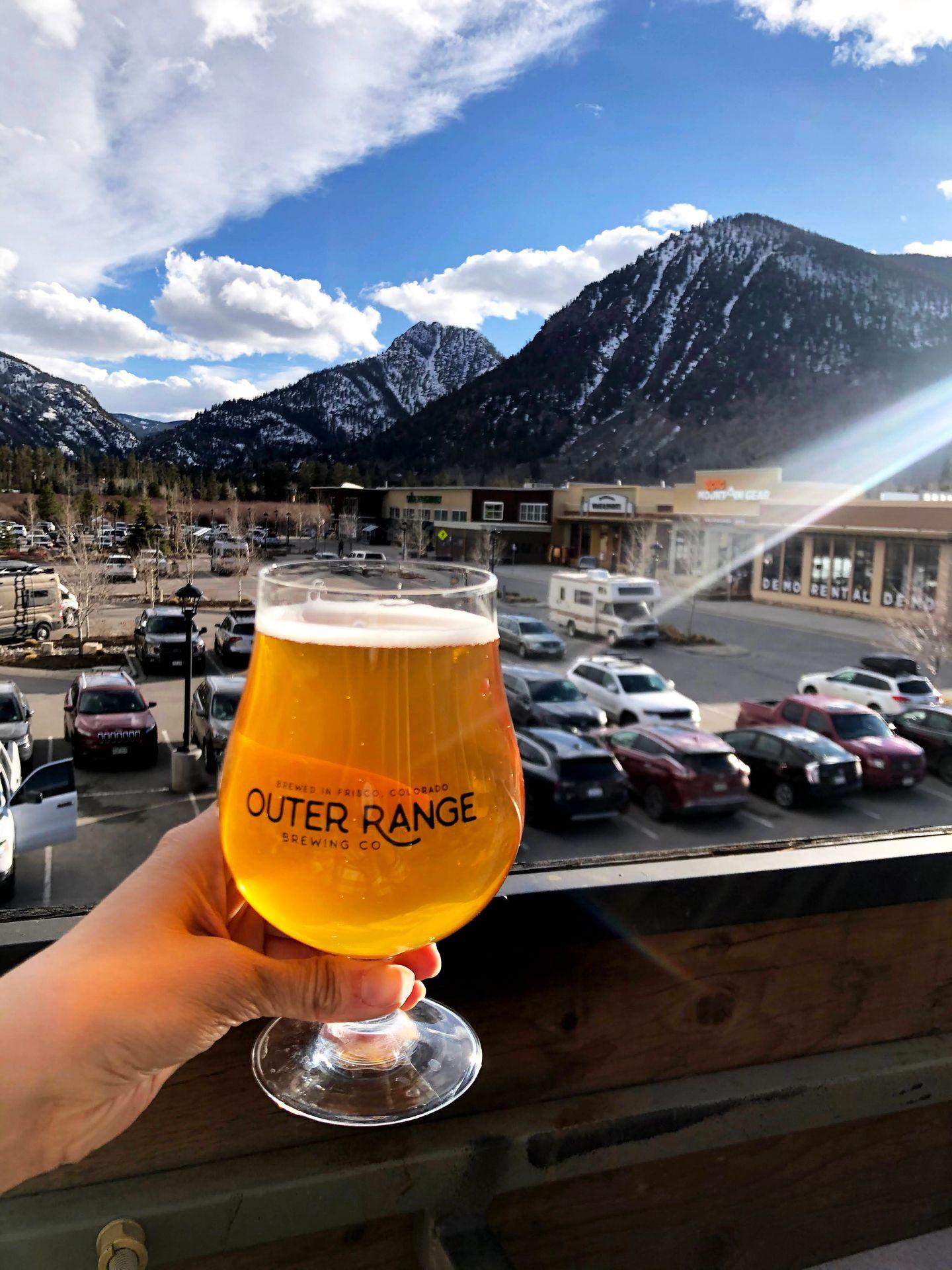Holding up a glass of golden beer on the deck of Outer Range. There is a snow covered mountain in the distance.