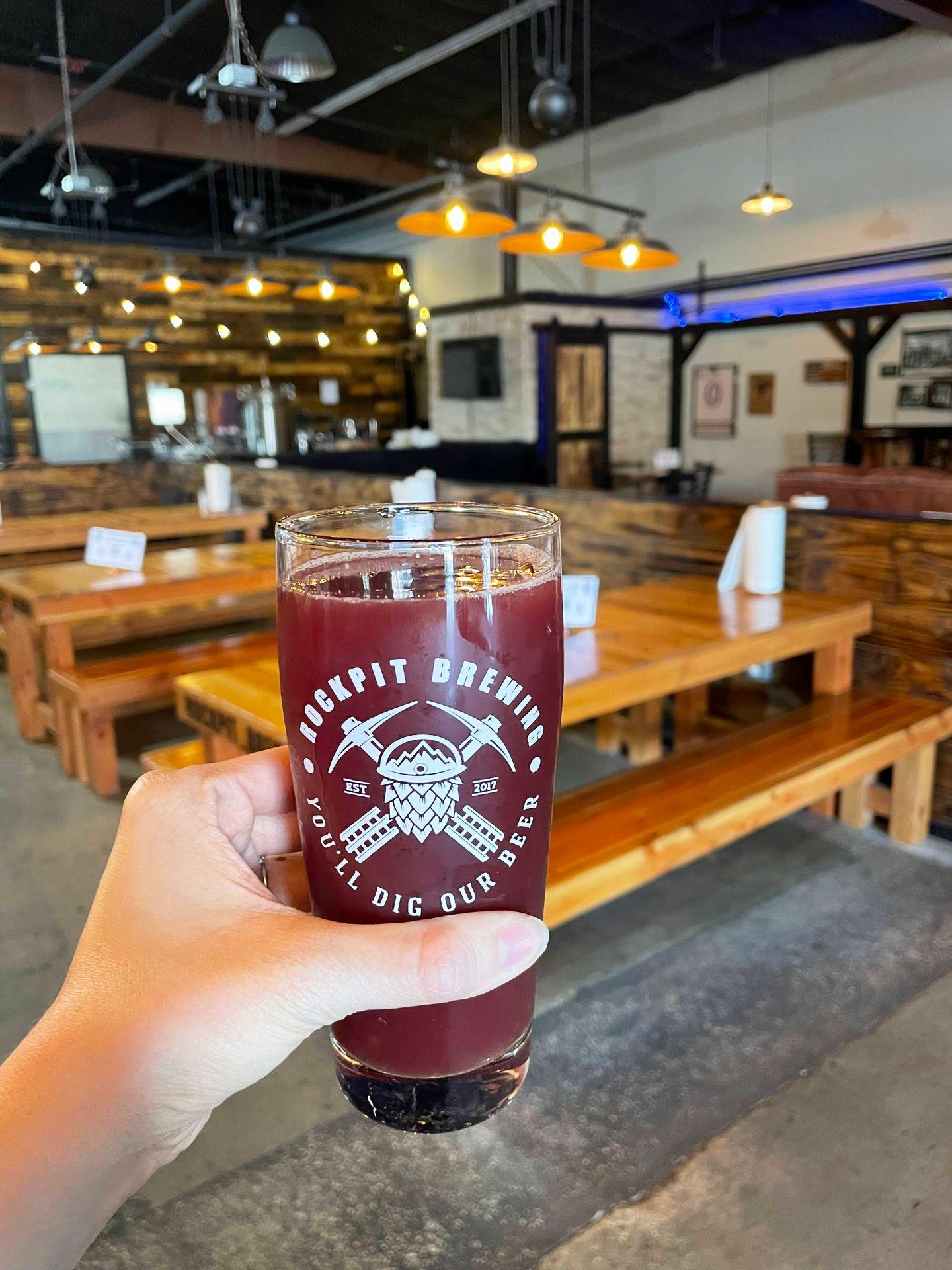 Holding up a smoothie sour beer inside of Rockpit Brewing. There are several long, wood tables in the background.