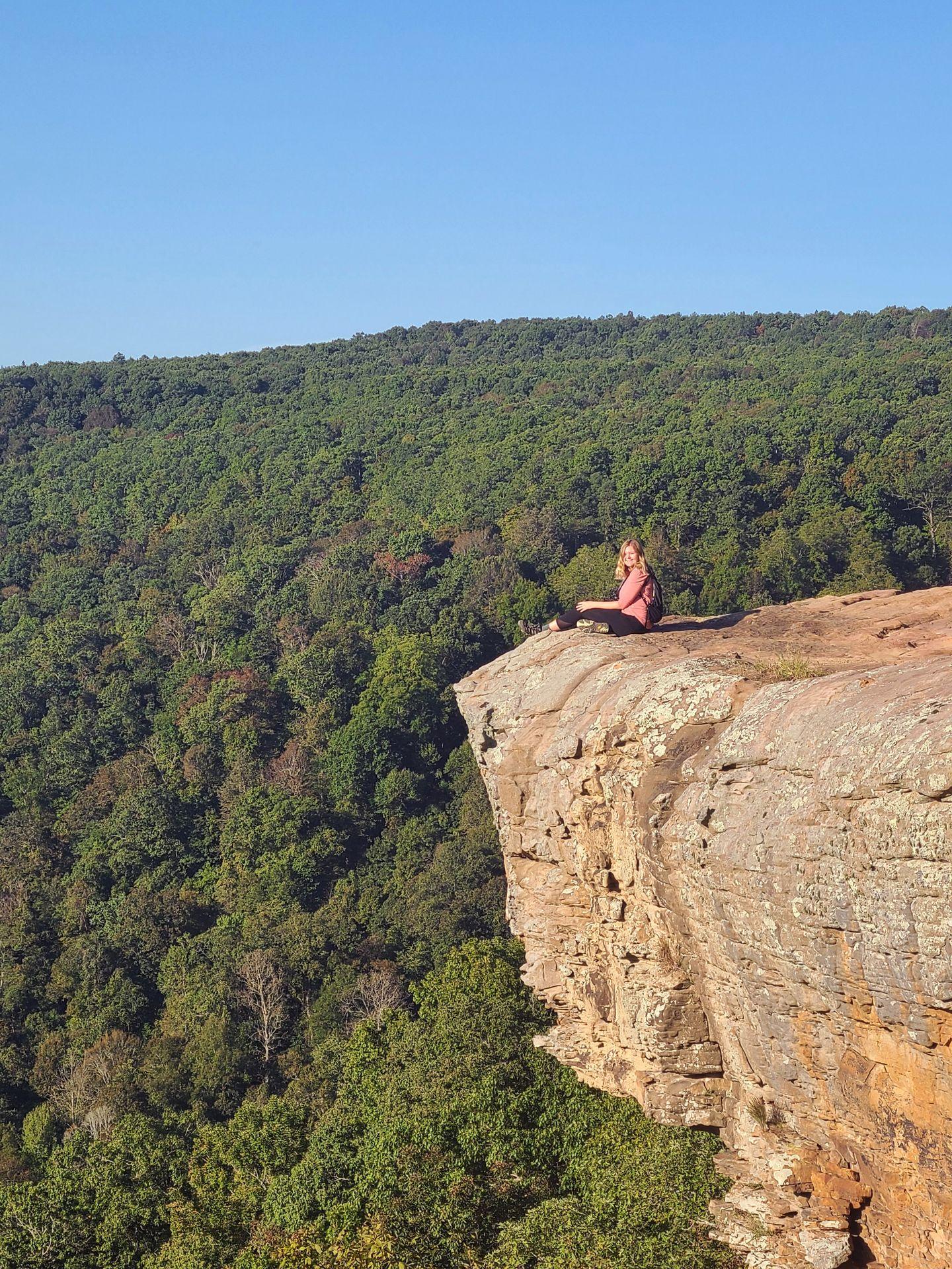 Lydia sits on the edge of the Whitaker Point rock with a view of green trees behind her.