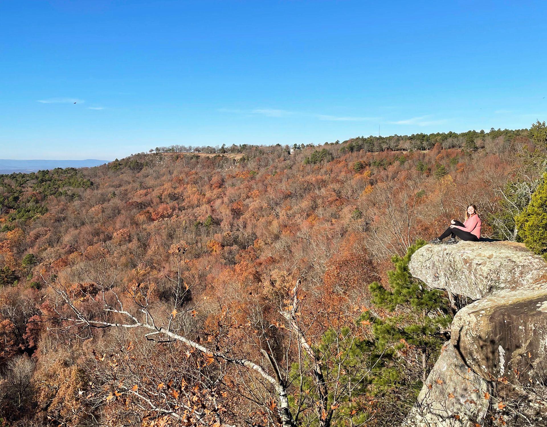 Lydia sitting on a rock that sticks out into the air. In the background, there is a mountain covered in trees with orange foliage.