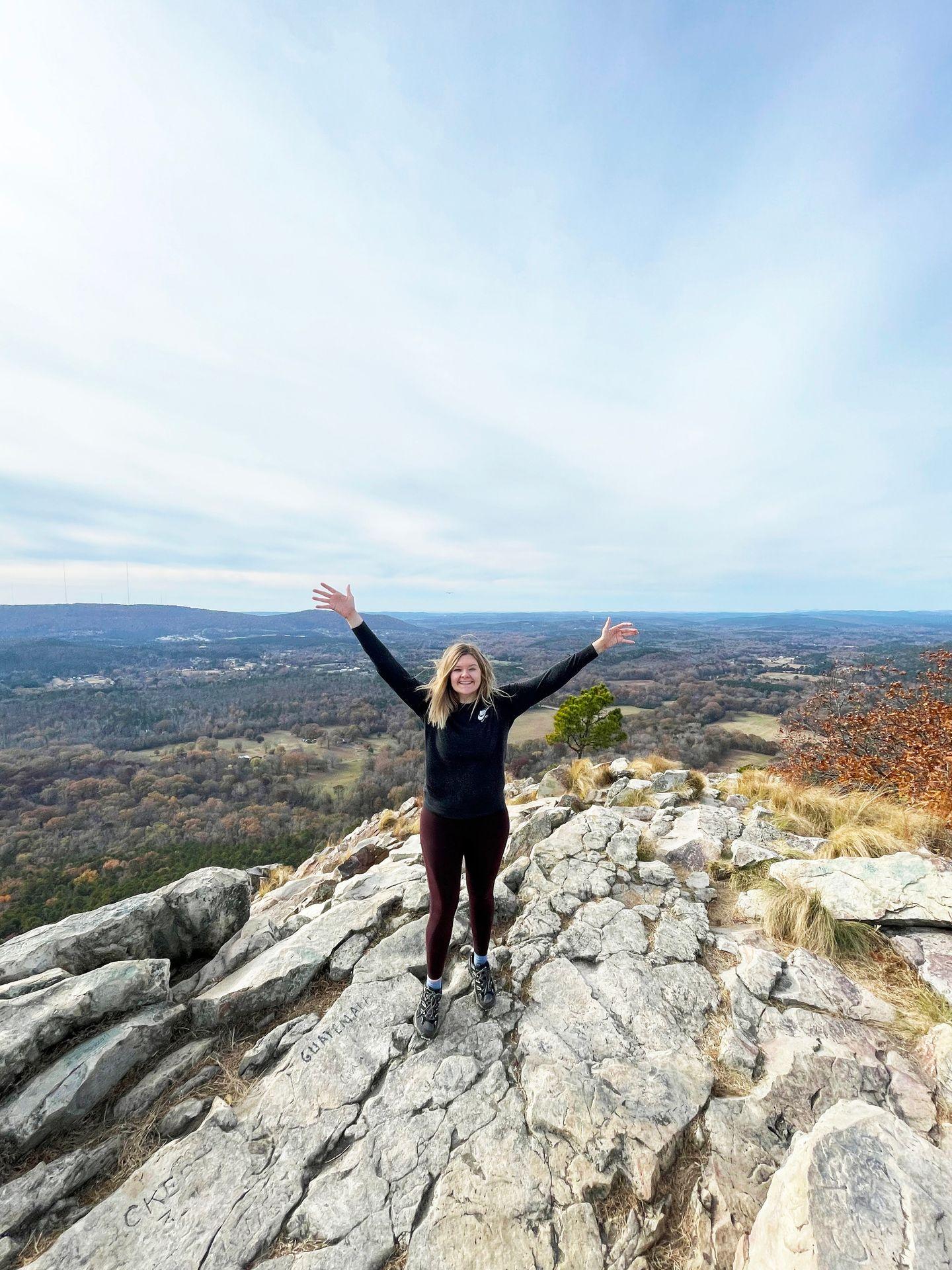 Lydia standing with her hands in the air on top of Pinnacle Mountain. She stands on white rocks with a view of trees in the background.