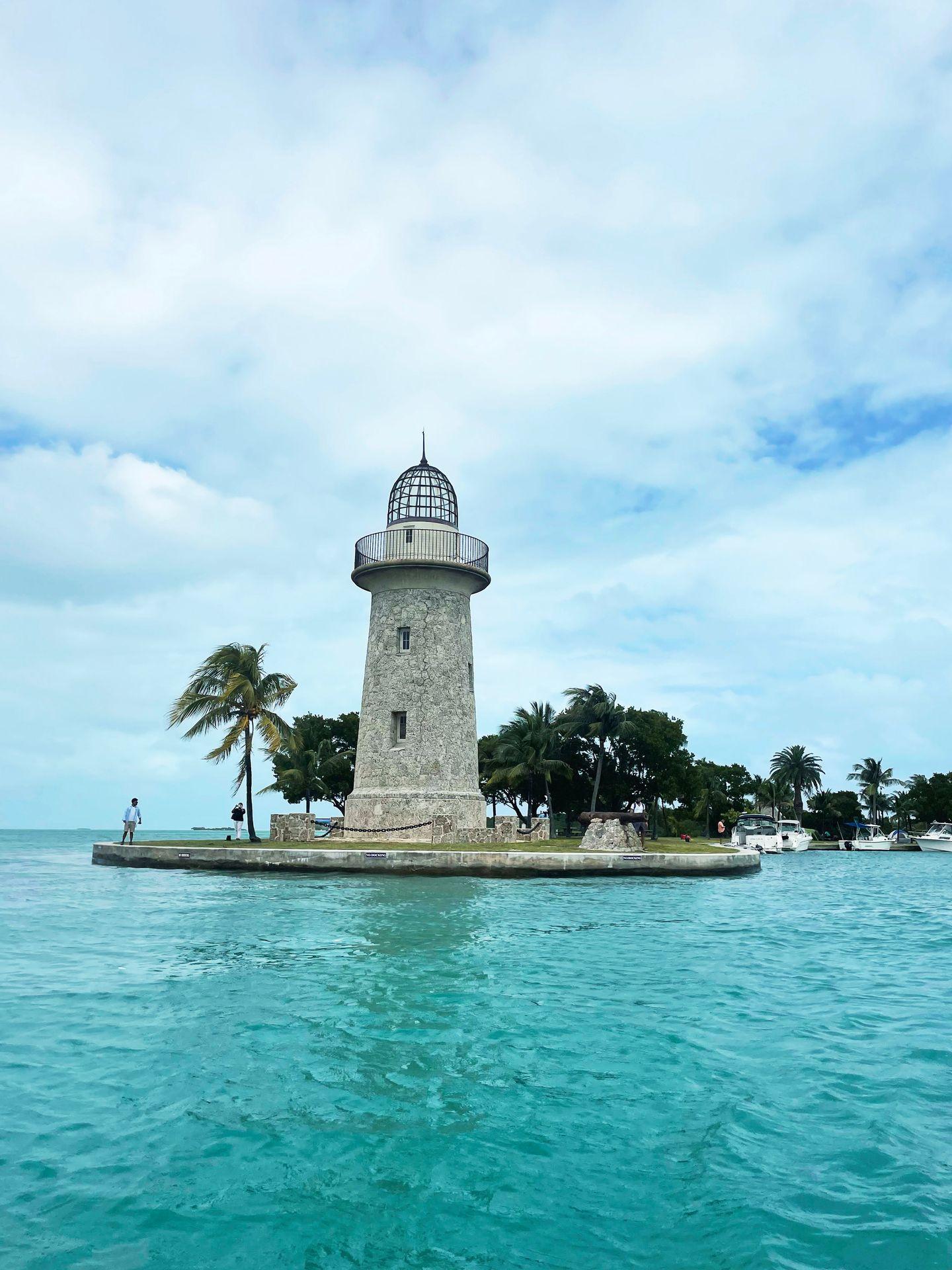 A view of the Boca Chita lighthouse from the water. The lighthouse is made of gray stone and there is a viewing balcony at the top. It is surrounded by palm trees and the ocean water in the foreground is aqua blue.