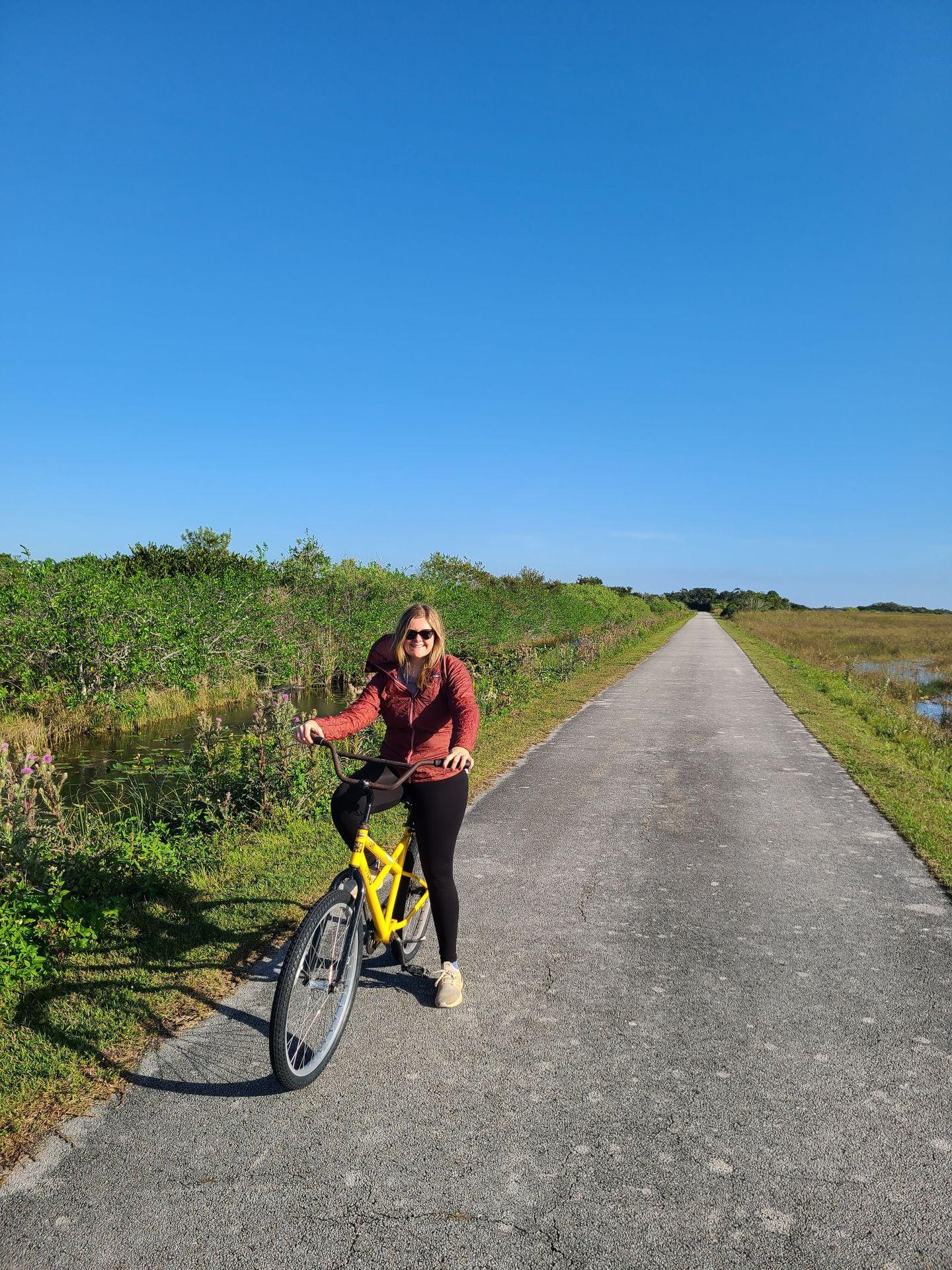 Lydia on a yellow bike on the Shark Valley bike trail. There is green grassland and a river on the side of the trail.
