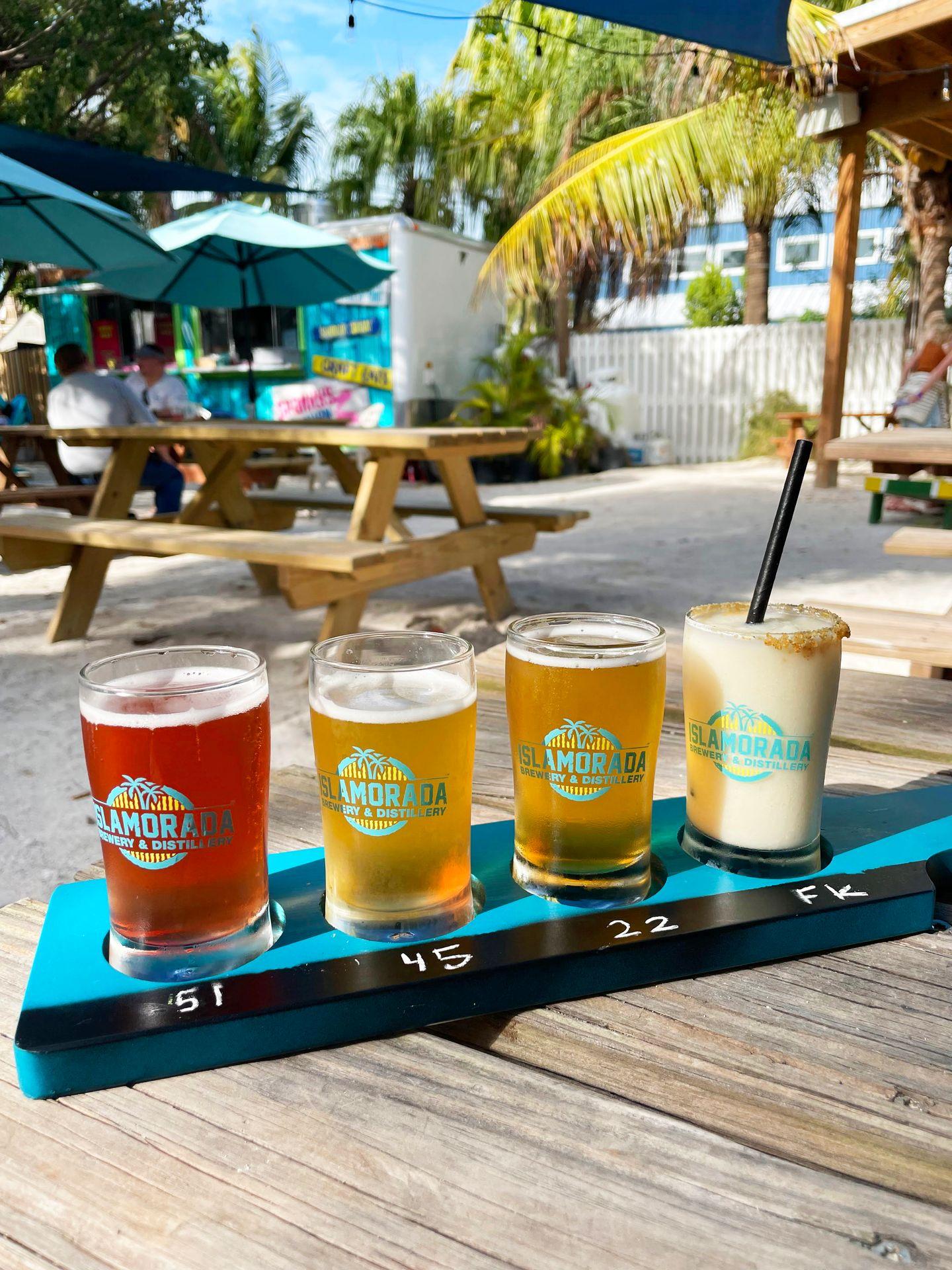 A flight of beer and a key lime pie cocktail at Islamorada Brewery. It sits on a picnic table in the outdoor patio area and there is a palm tree in the background.