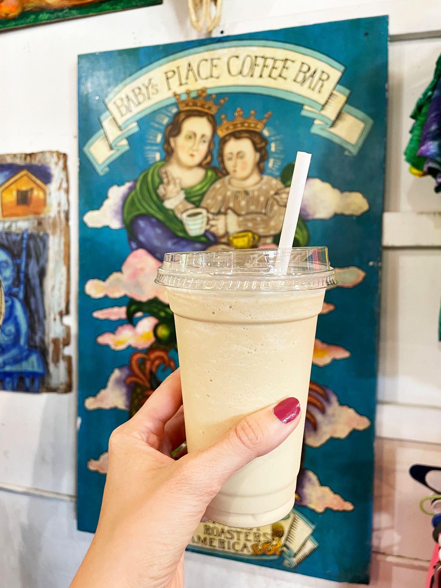 A frozen latte being held up in front a painting with angels with the words 'Baby's Place Coffee Bar in the background