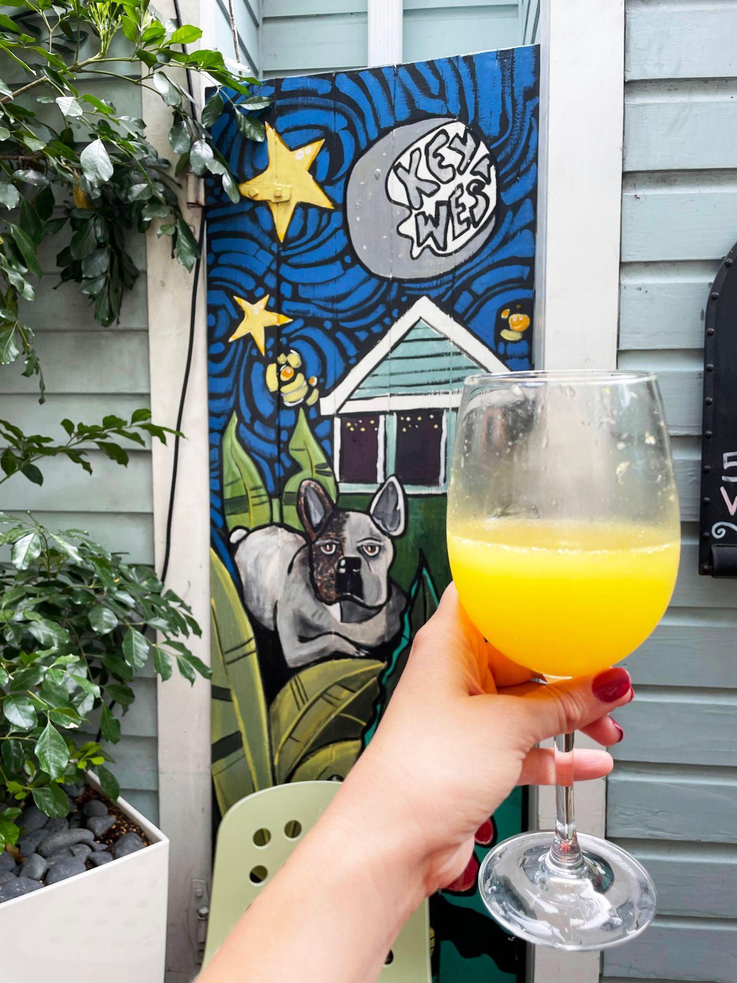 A mimosa being held up in front of a mural with a dog, a house and the words Key West in the sky