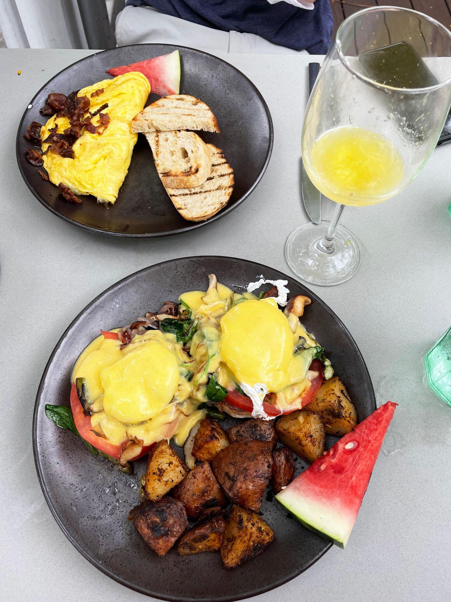 Two breakfast plates and a mimosa. One is caprese eggs benedict and the other is an omelette with toast and watermelon