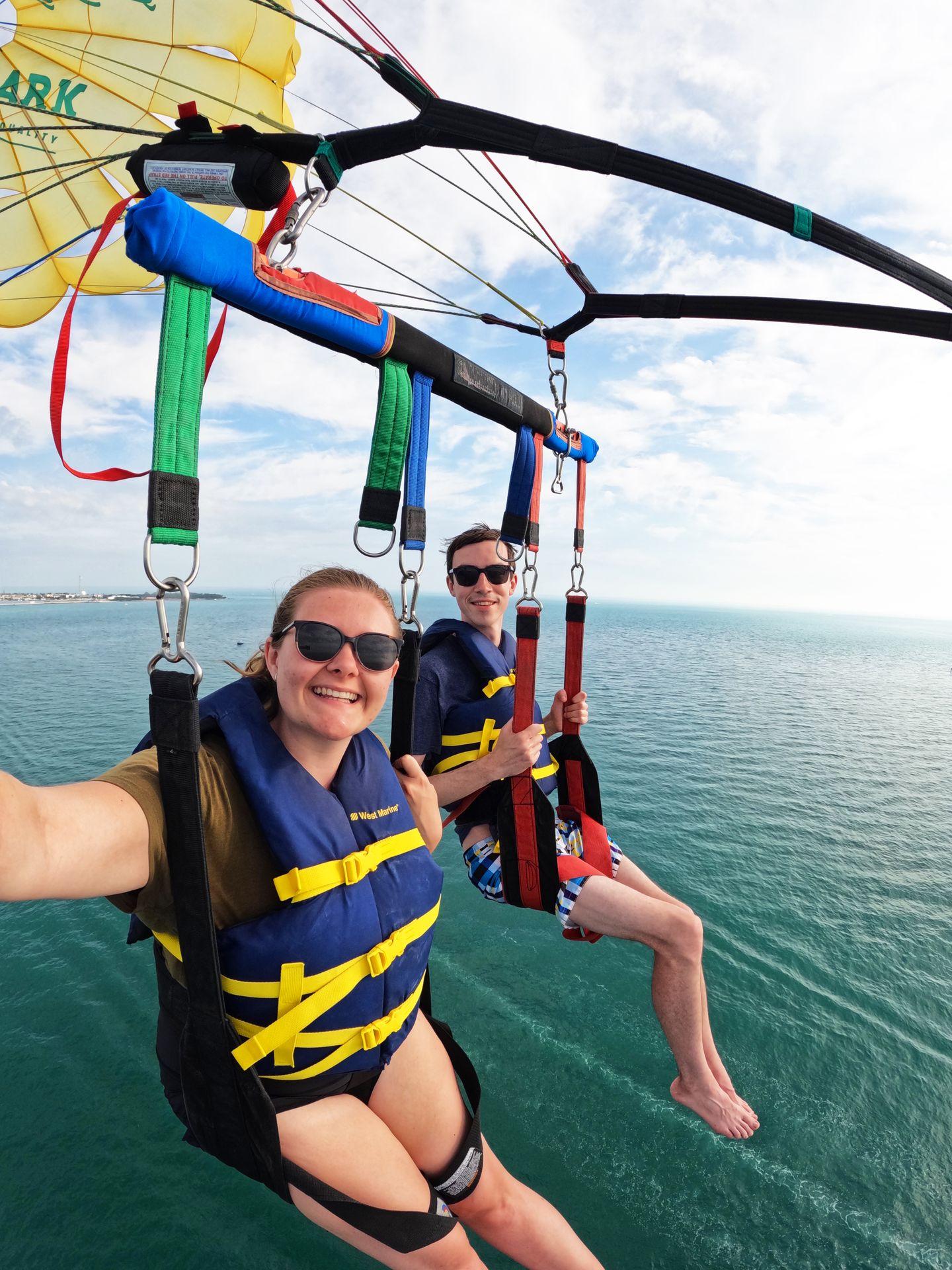Lydia and Joe sitting up in the sky while parasailing. They are harnessed in and wearing life jackets