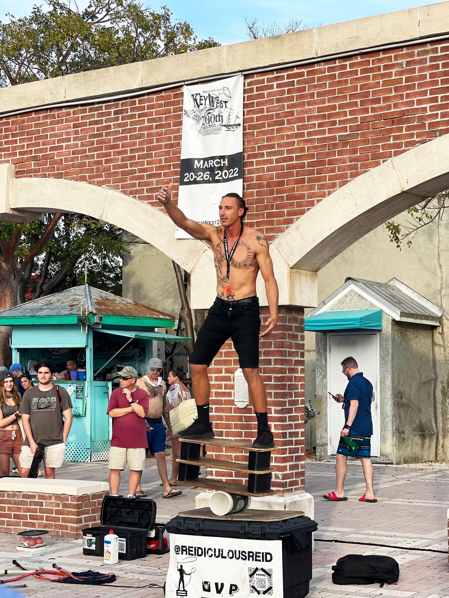 A street performer balancing on a wooden board on top of a cylinder. The man is shirtless with chest tattoos