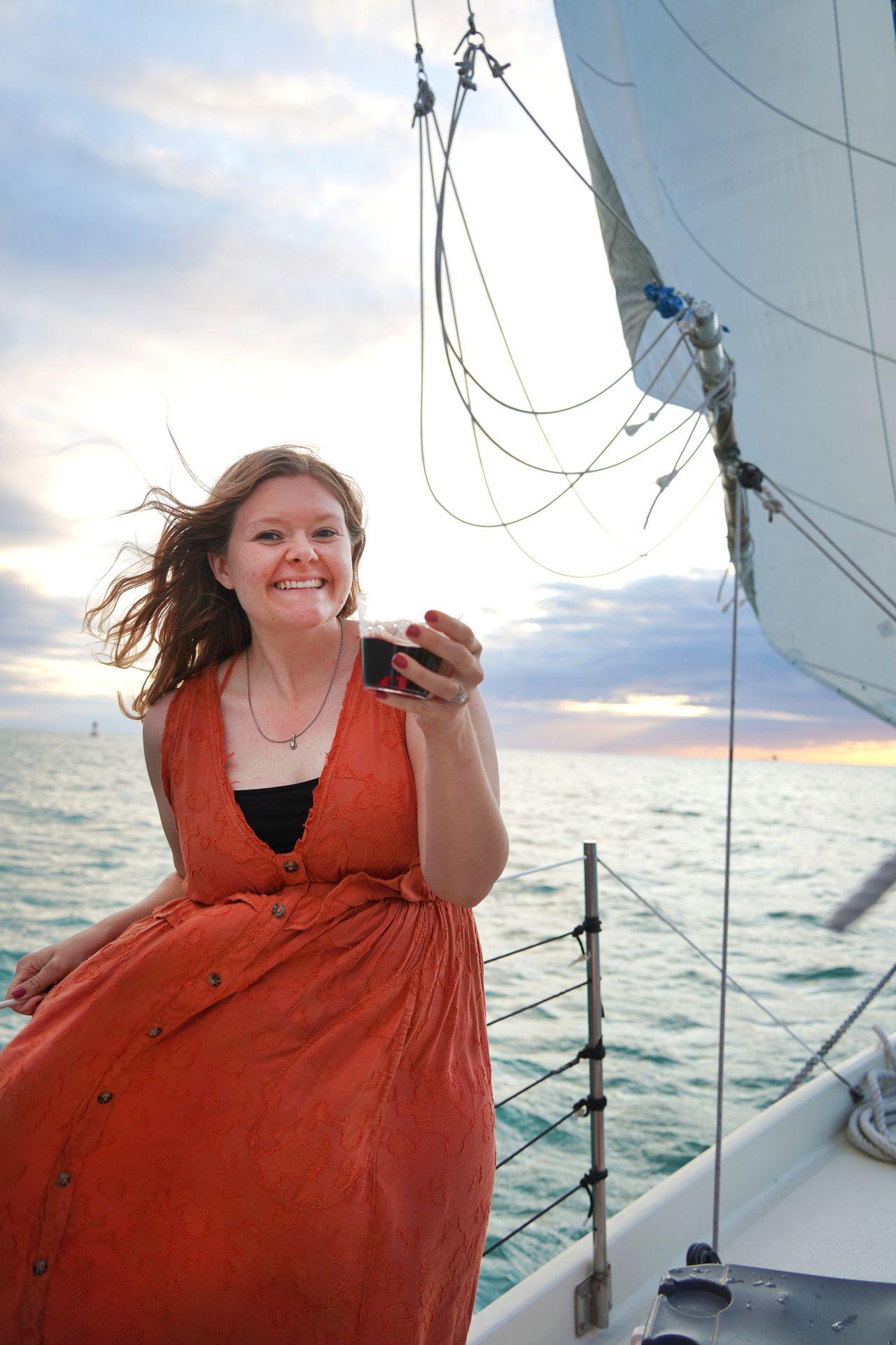 Lydia on a sailboat holding a cup of red wine. Her dress and hair blows in the wind