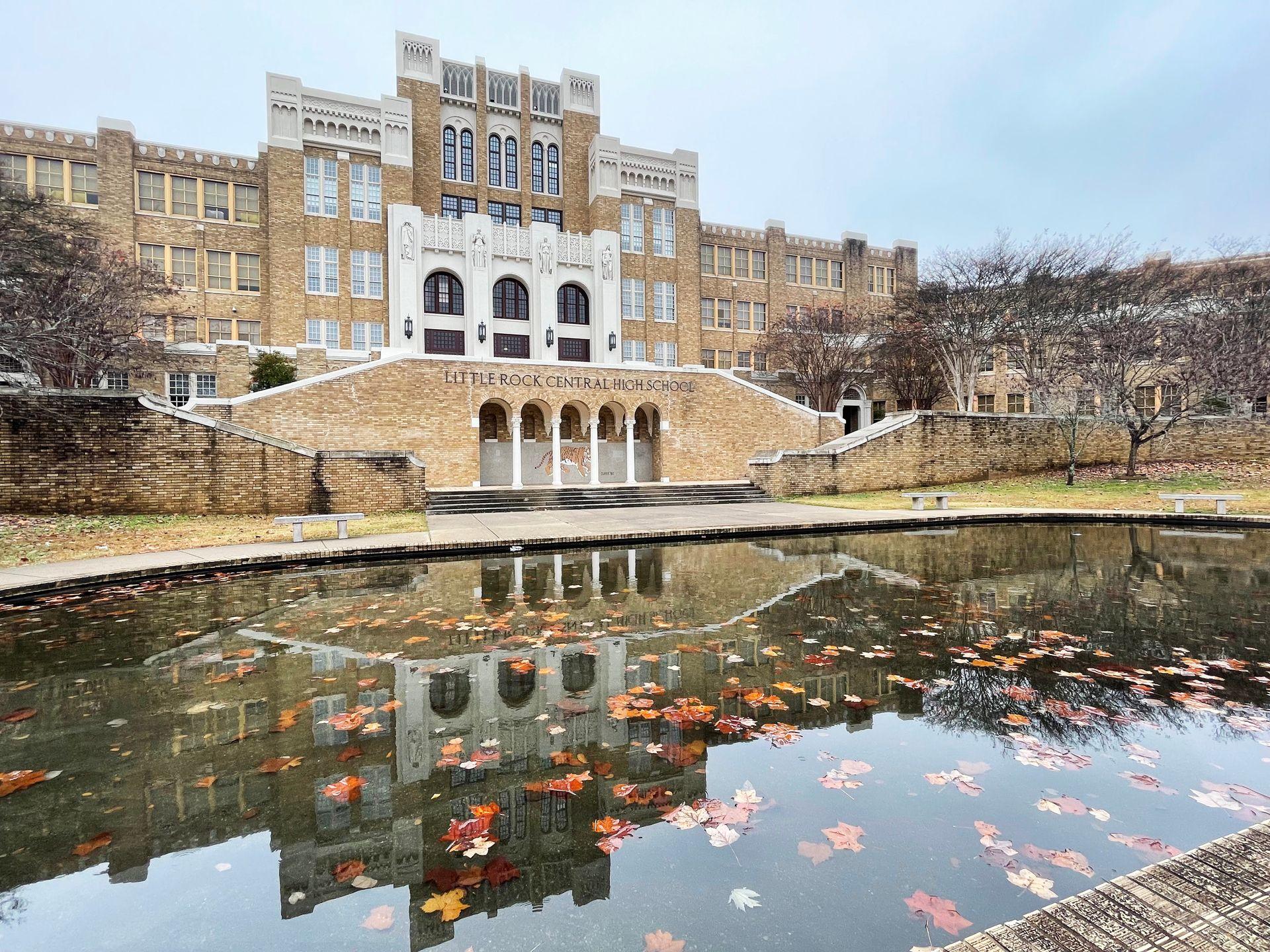 The exterior of Little Rock Central High School. The school reflects in a pool of water with some fall leaves floating in the water.