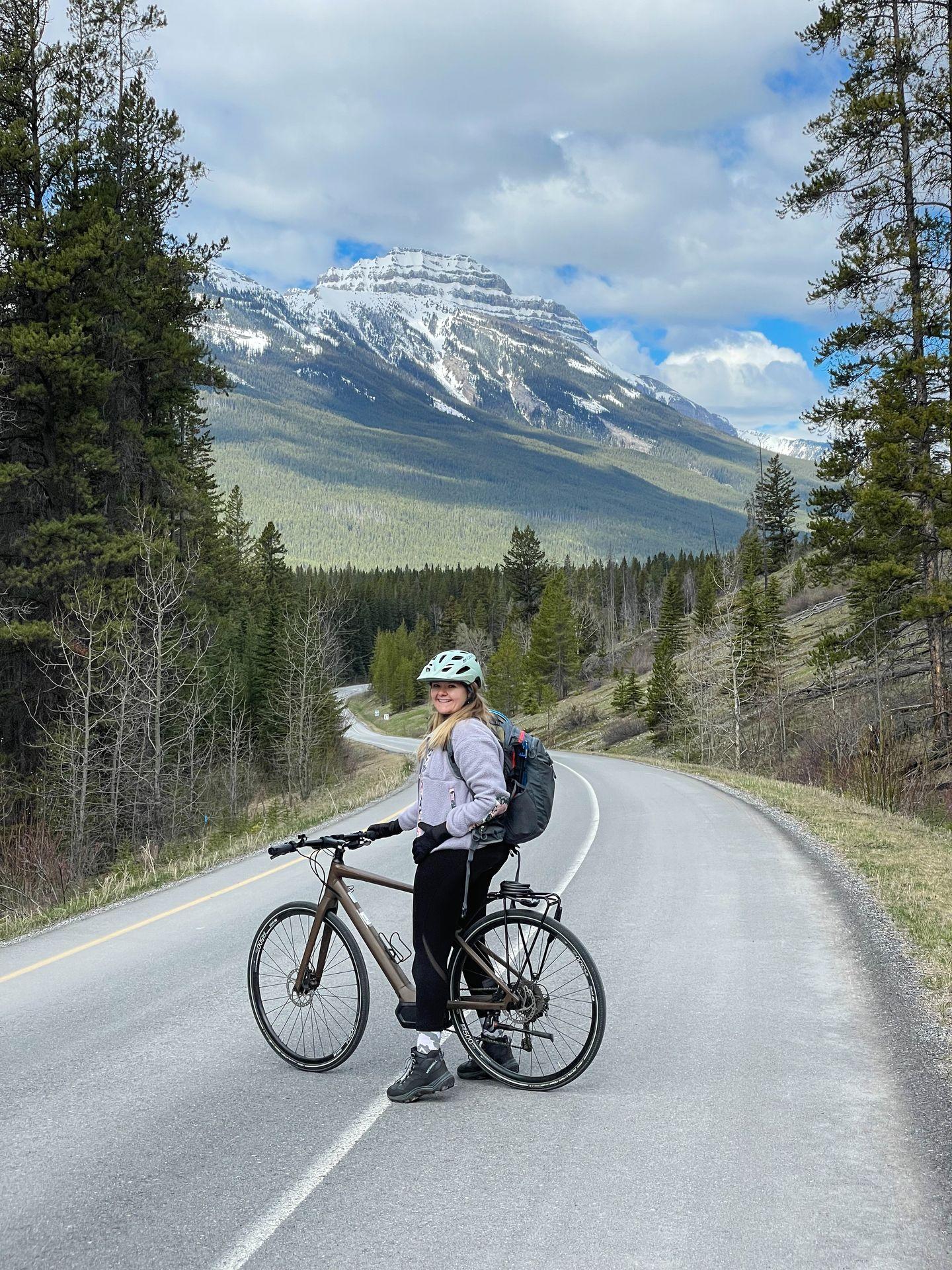 Lydia on a bike on the Bow Valley Parkway. The road curves downward and there is a tall mountain in the background.