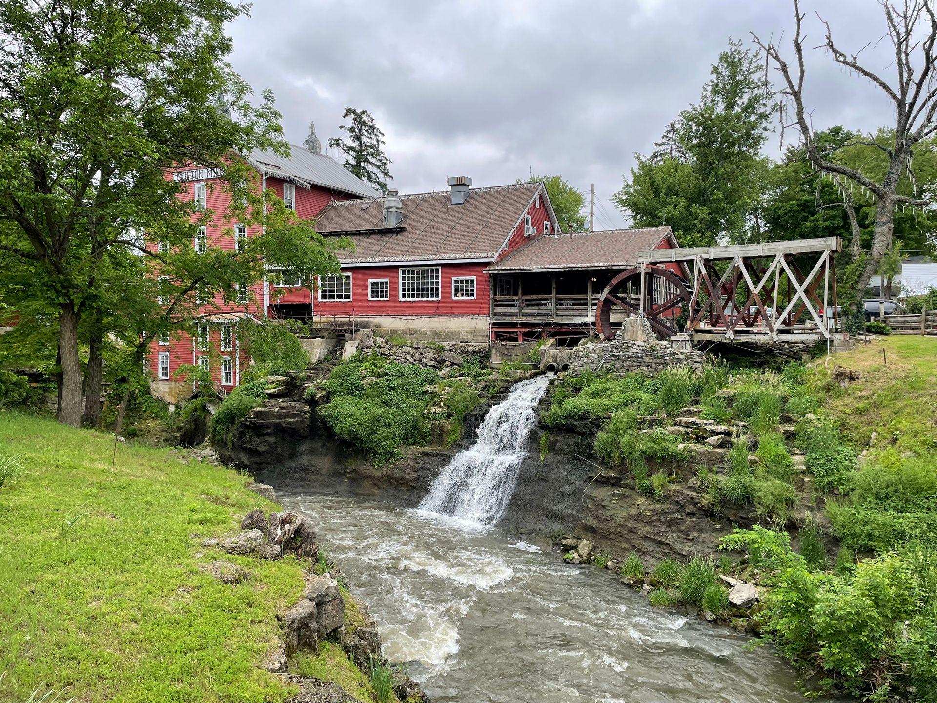 Looking at Clifton Mill. The building is red and has a water mill. Water flows down from the Mill and into a river.