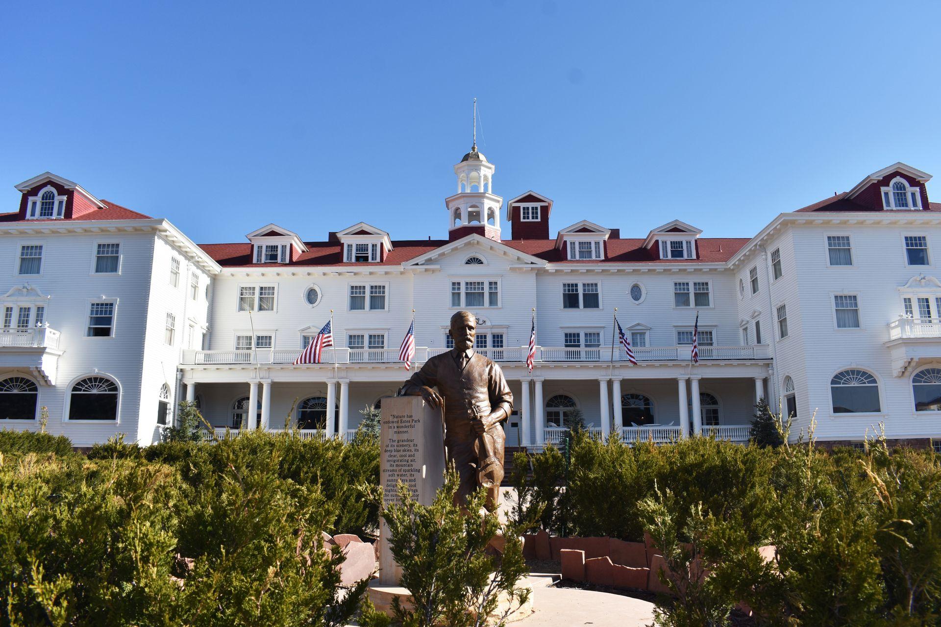 The exterior of the famous Stanley Hotel. The hotel is white with a red roof and has a large porch. There is a statue of a man leading on a pillar directly in front of the hotel.