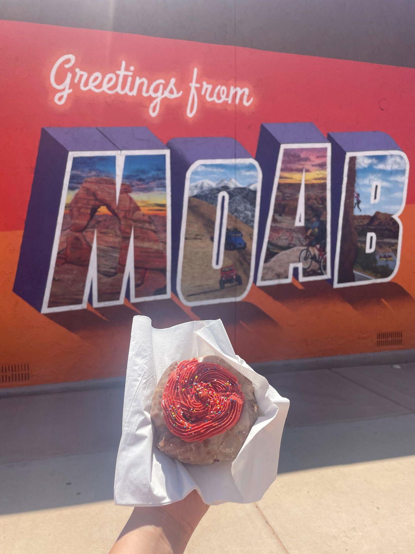 Holding up a donut with red icing in front of a "Greetings from Moab" mural