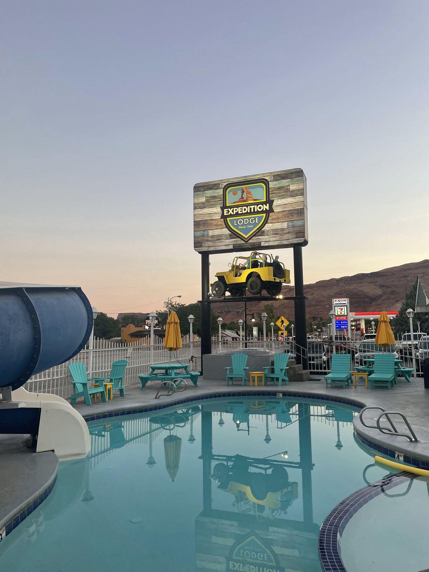A pool surrounded by blue chairs. In the background, there is a sign for Expedition Lodge with a yellow Jeep up in the air.