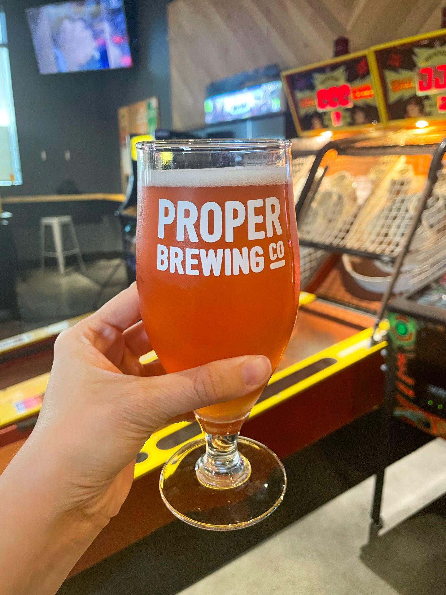 Holding up a beer in front of a skee-ball game in Proper Brewing.