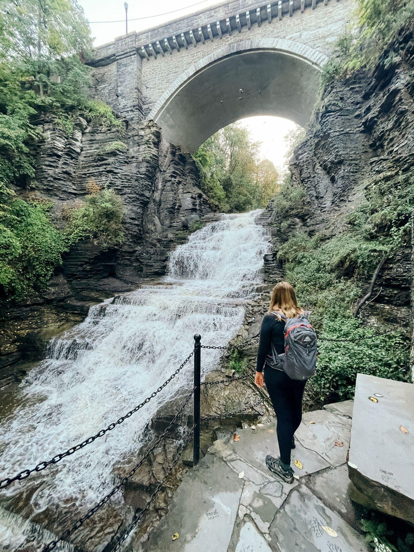 Lydia standing and looking out at a waterfall on the Cascadilla Gorge Trail. There is a stone bridge above the waterfall.