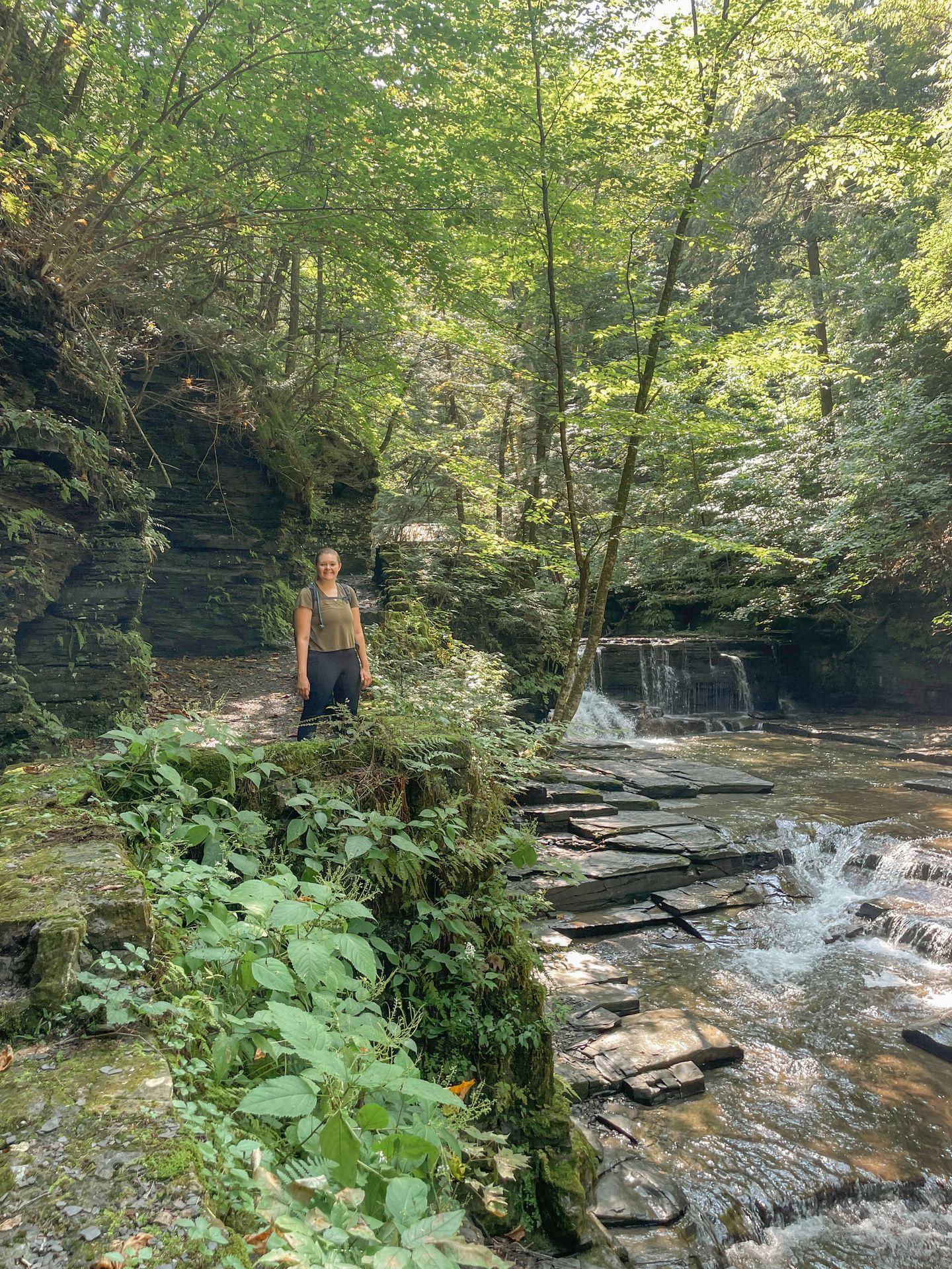 Lydia standing on a trail in Fillmore Glen. The path is covered in green moss and vines and there is a stream with a waterfall to the right.