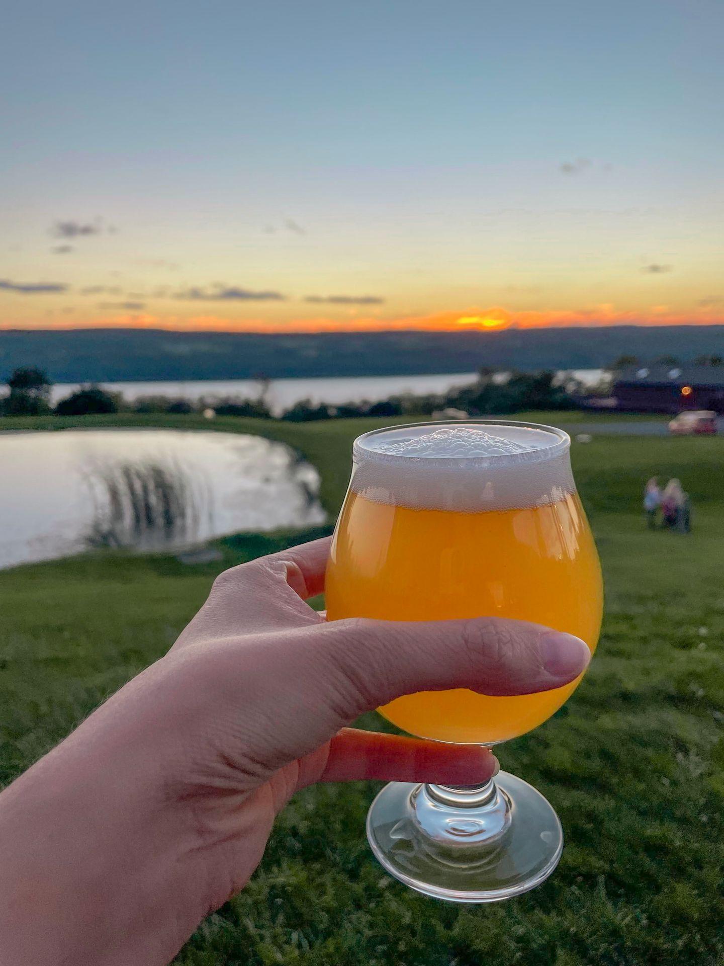 Holding up a glass of beer with a pond, a lake and the setting sun in the distance.