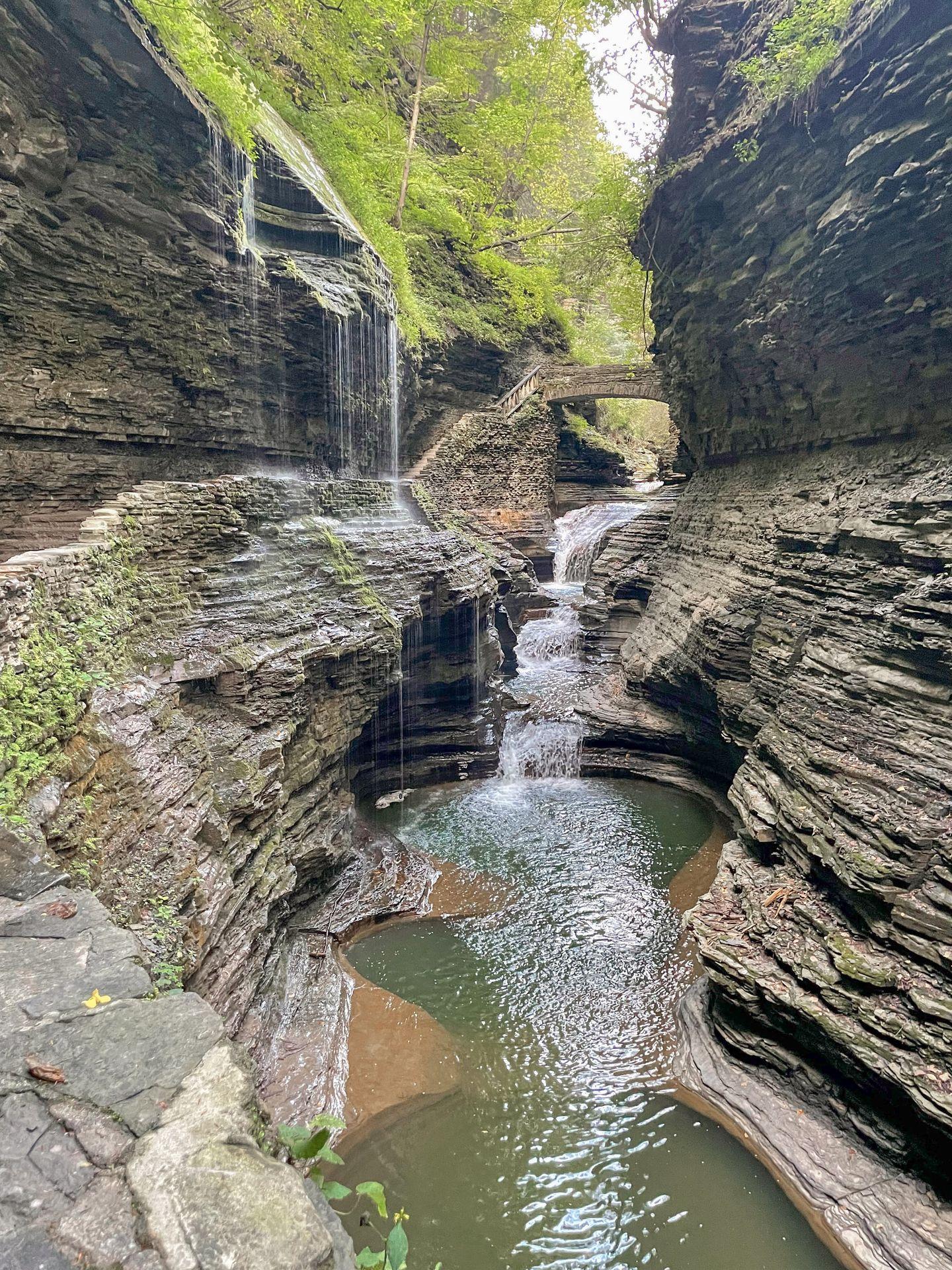 A view of the gorge at Watkins Glen that includes a waterfall, a stone bridge and water falling from the left side of the top of the gorge.