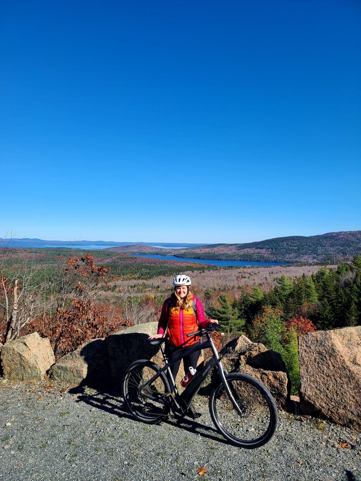 Lydia standing with her bike with a view of a lake in the background from a Carriage Road in Acadia.