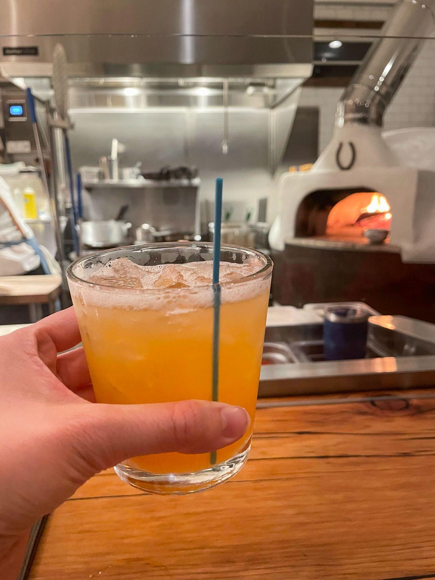 Holding up a cocktail with a wood-fired pizza oven in the background.