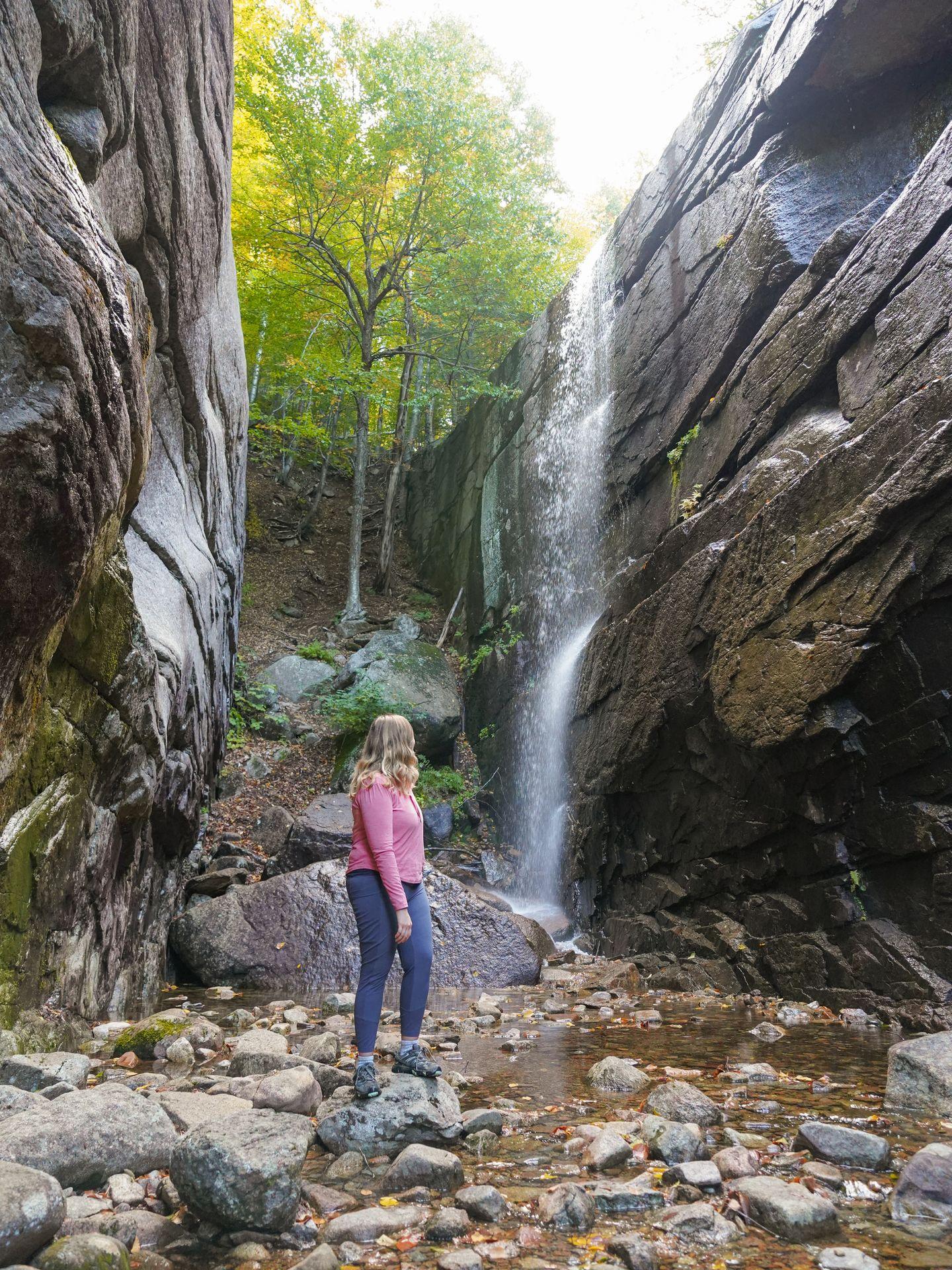 Lydia standing between two tall, flat rock cliffs. A waterfall flows over the side of one of the rocks.
