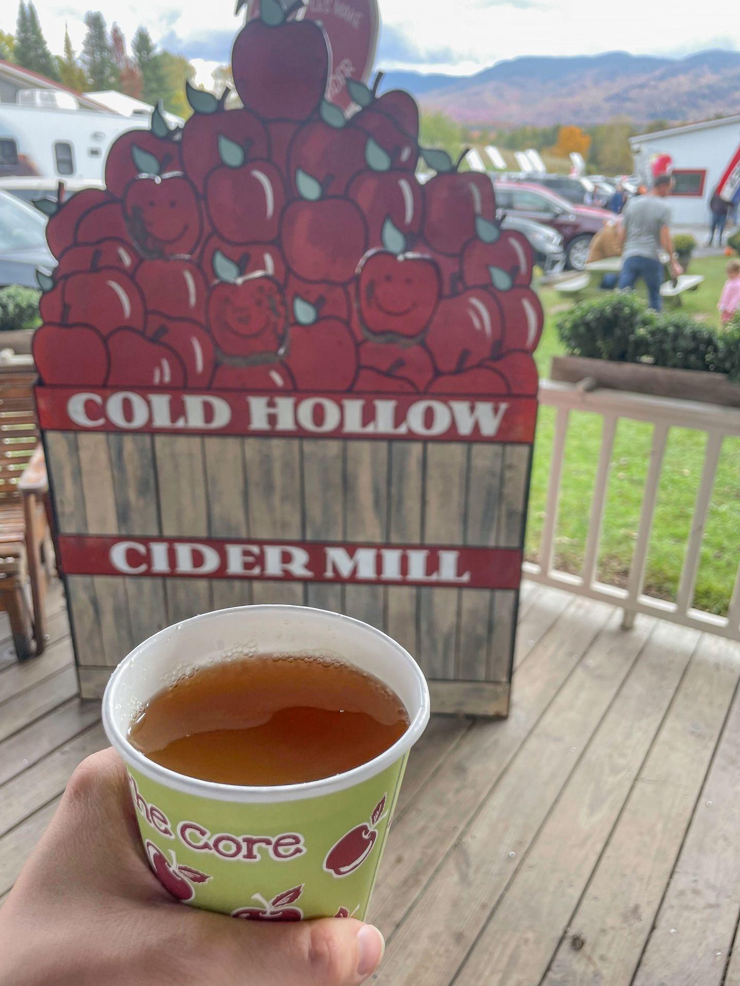Holding a cup of hot apple cider in front of a wooden sign that reads 'Cold Hollow Cider Mill'
