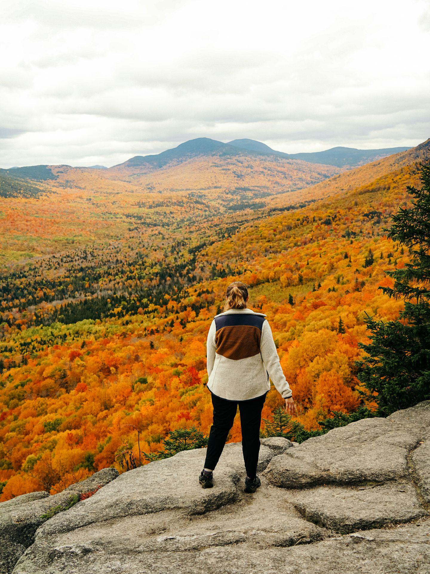 Lydia standing on a cliff and looking out a view of bright orange leaves.