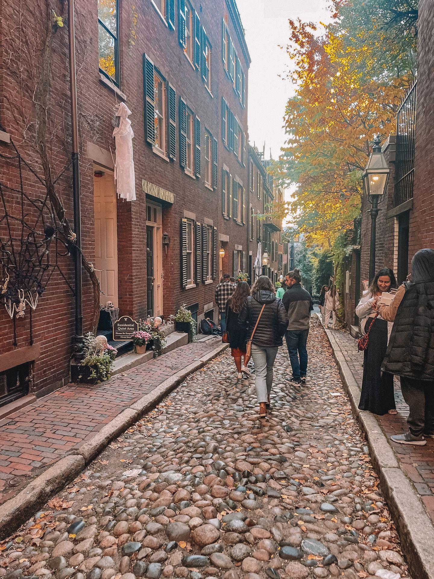 A crowded Acorn Street, which is made of cobblestone. There is Halloween decor on some of the houses.