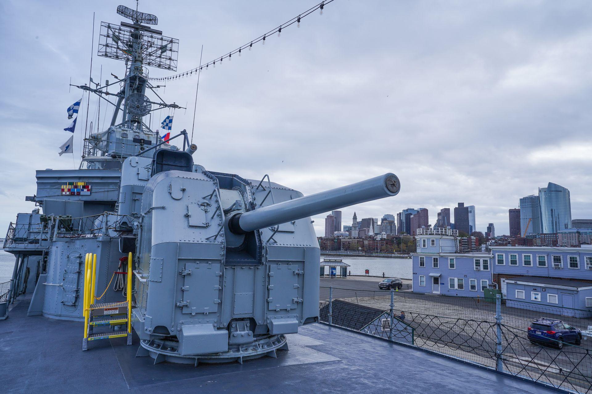 A weapon on top of the USS Cassin Young. The ship floats in the harbor and you can see the downtown Boston in the background.