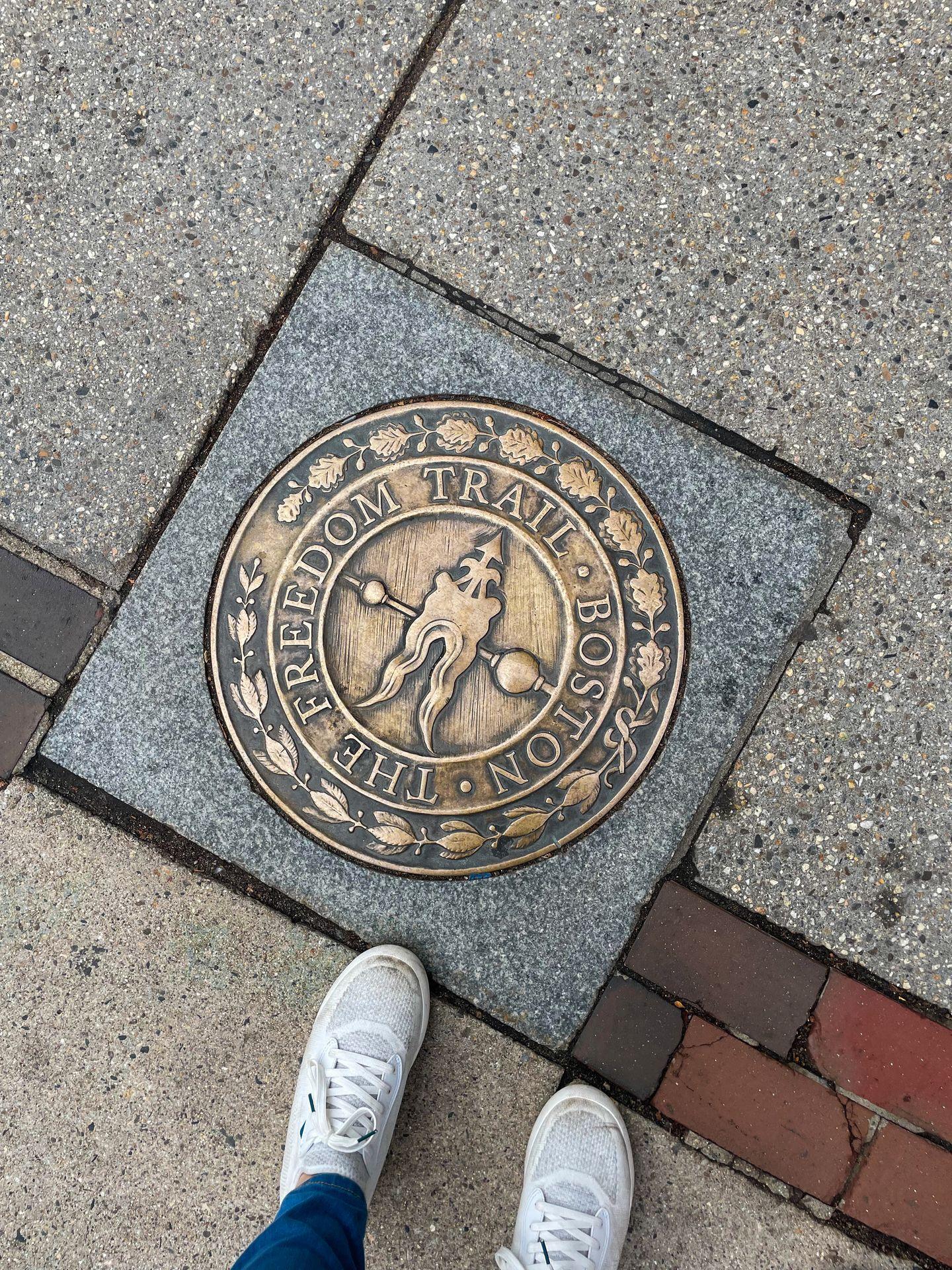 A plague on the ground that marks the Freedom Trail in Boston. The label is gold and Lydia's feet are next to the sign.