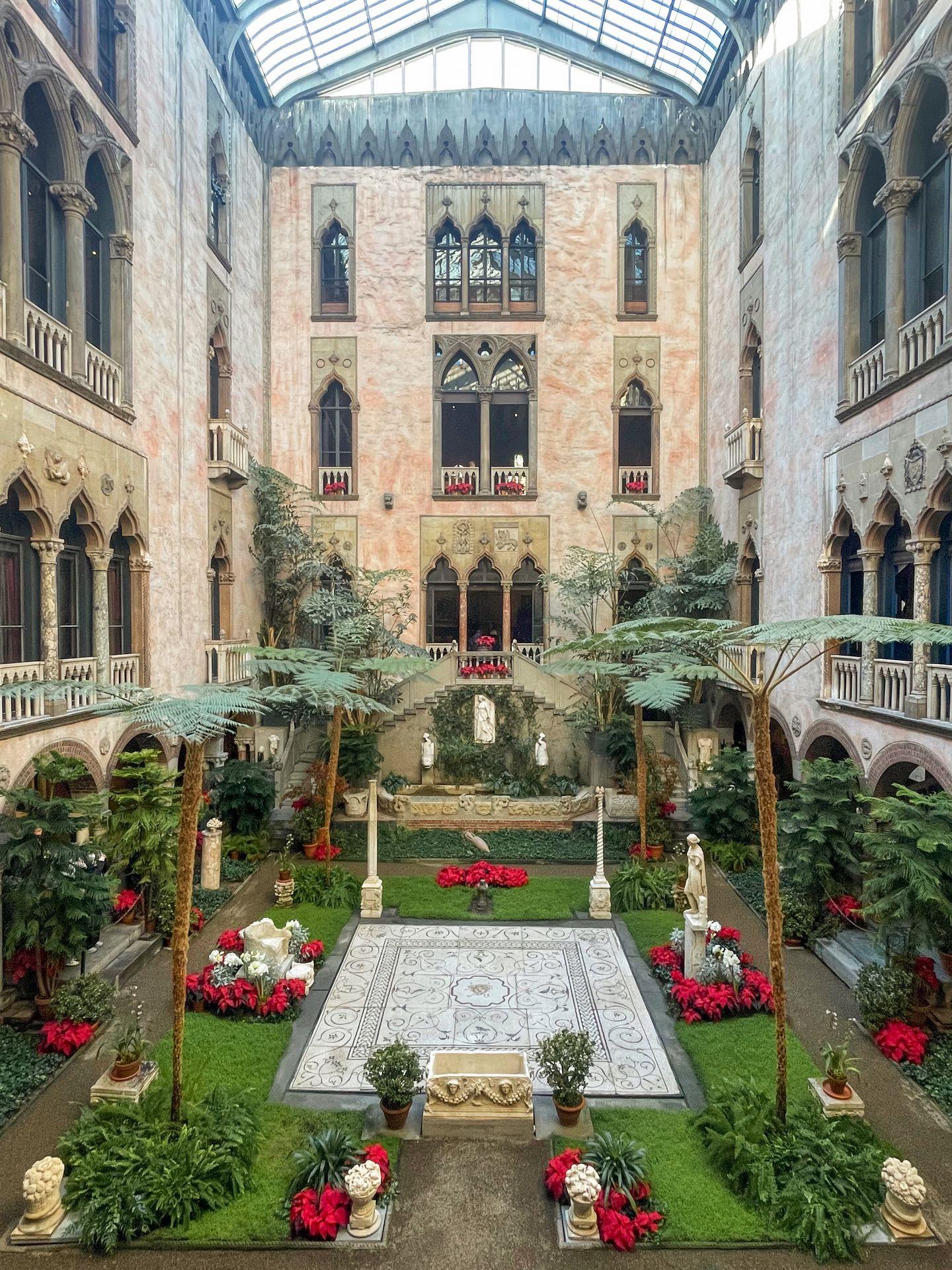The courtyard in the center of the Gardner Museum. Several stories of the building surround the courtyard. There is a tile floor in the center of the ground that is surrounded by plants. There are red flowers and greenery.