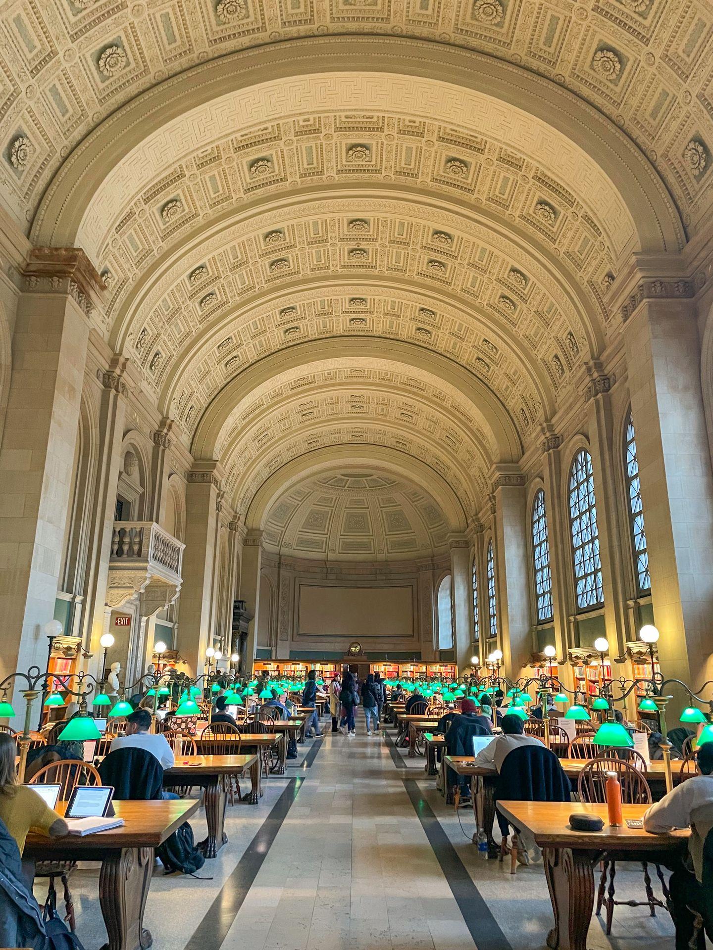 Inside of the Bates Hall in the Boston Public Library. There are several tables with green lamps and a tall, arched ceiling