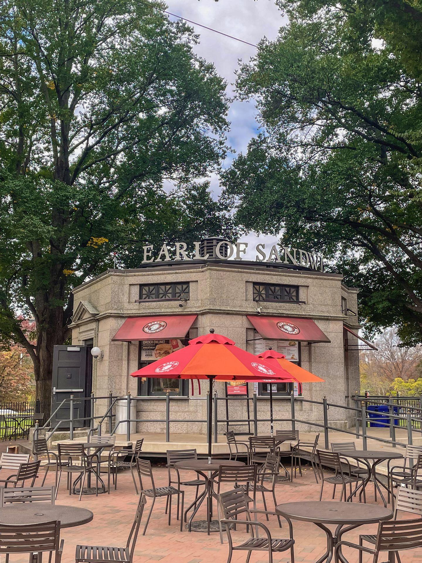 The Earl of Sandwich restaurant inside of Boston Common. There are tables outside of hte building with red and orange umbrellas.