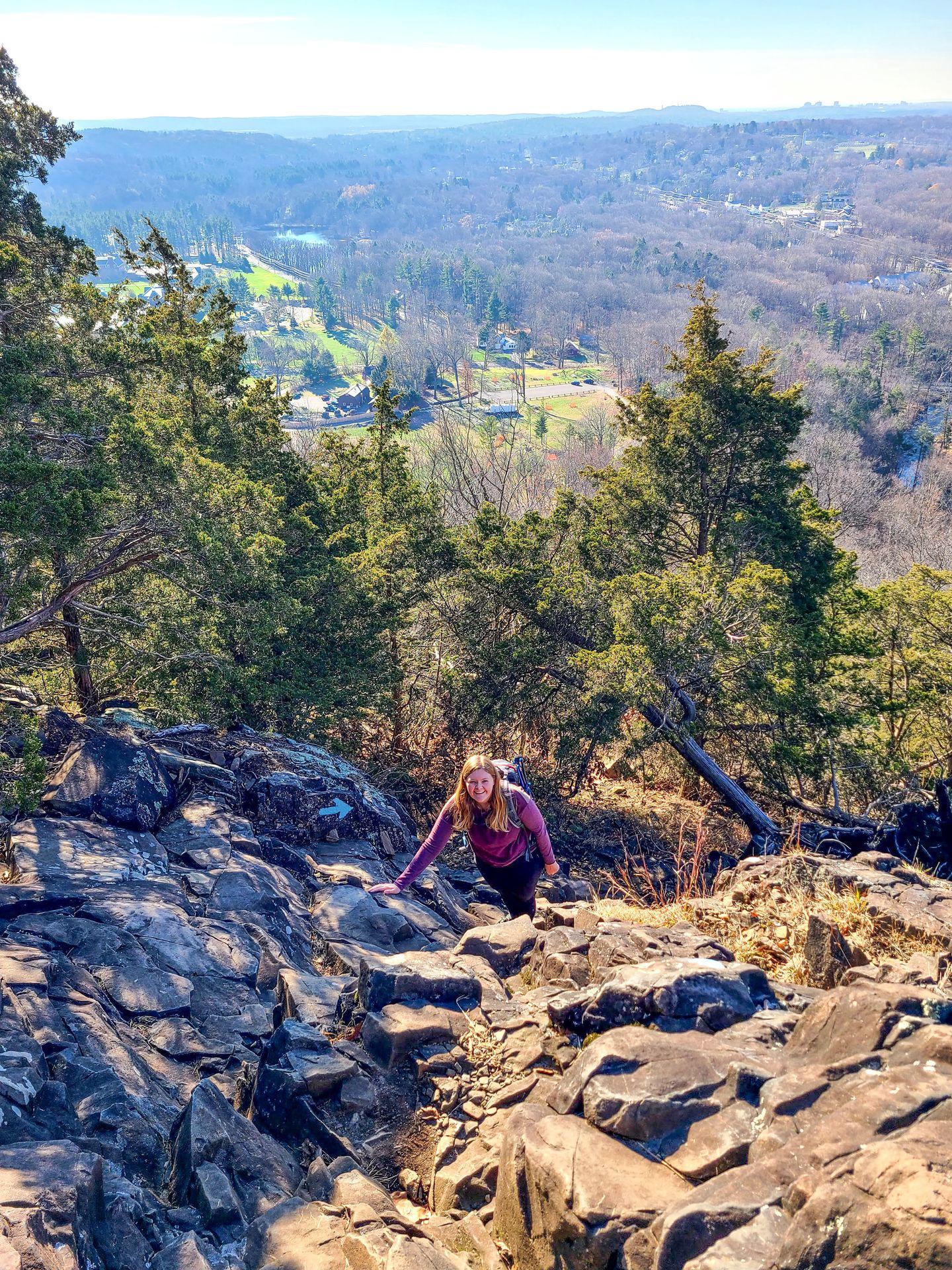 Lydia scrambling up the blue trail in Sleeping Giant State Park. There is a view of a valley down below in the background.