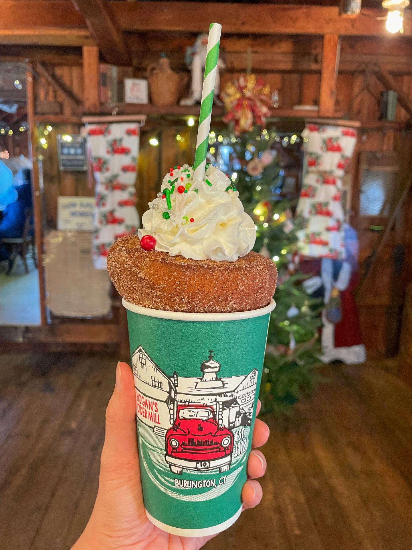 A cup of hot cider topped with an apple cider donut and whipped cream. The cider is in a green, paper cup with artwork depicting Hogan's Cider Mill.