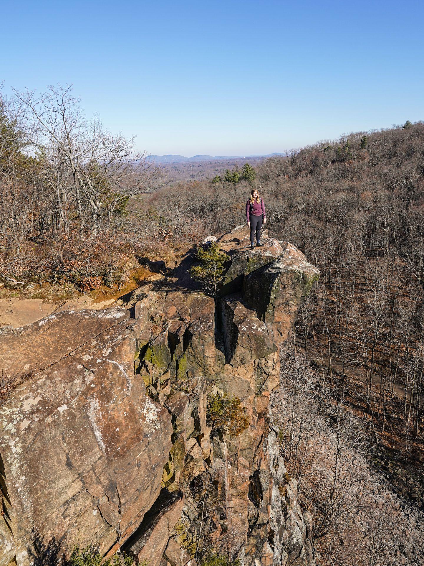 Lydia standing out on a large rock formation with a steep cliff on one side in Sleeping Giant State Park.