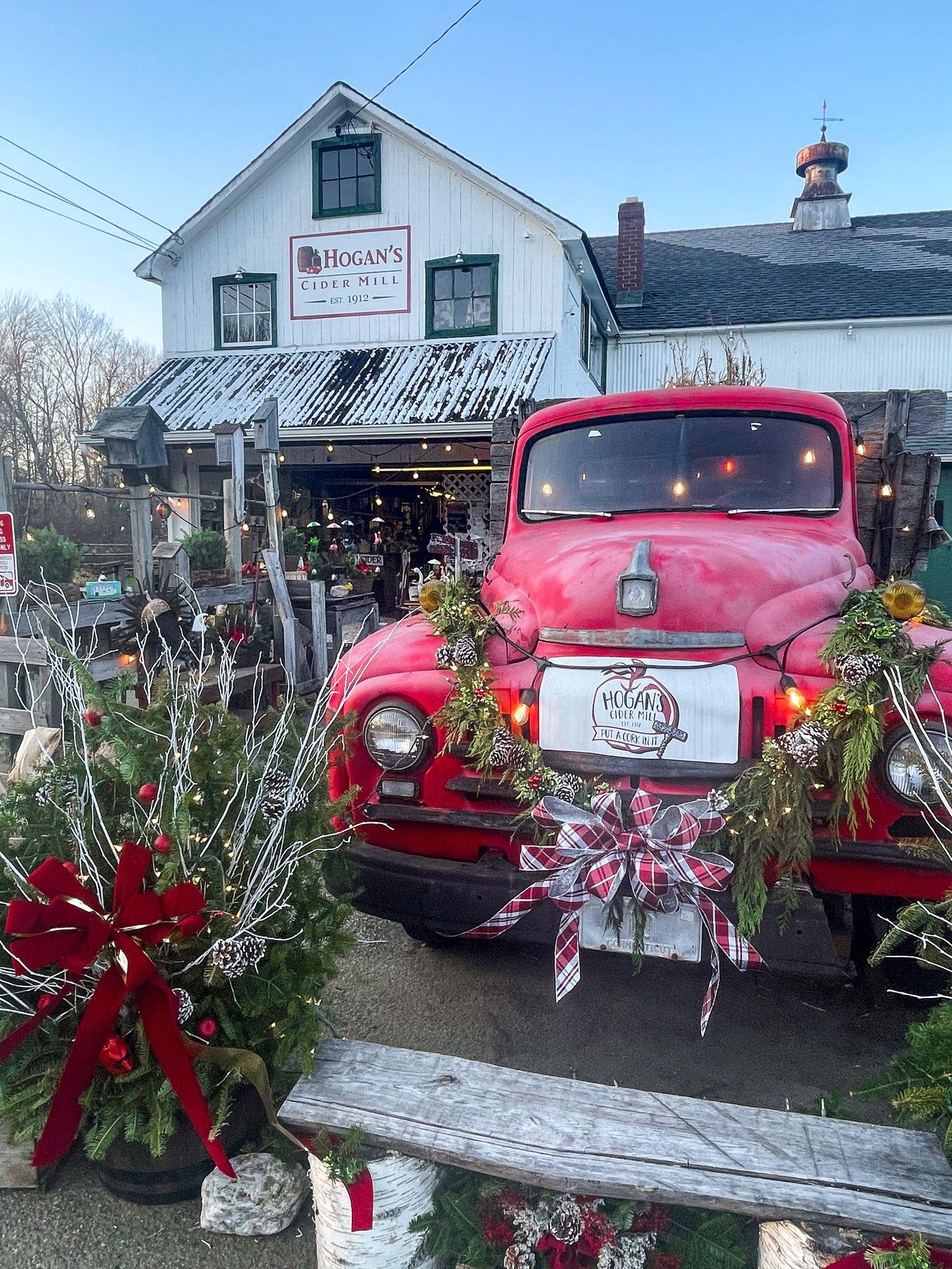 The exterior of Hogan's Cider Mill. There is a vintage red truck adorned with garland and a red bow. The general store in the background is white.