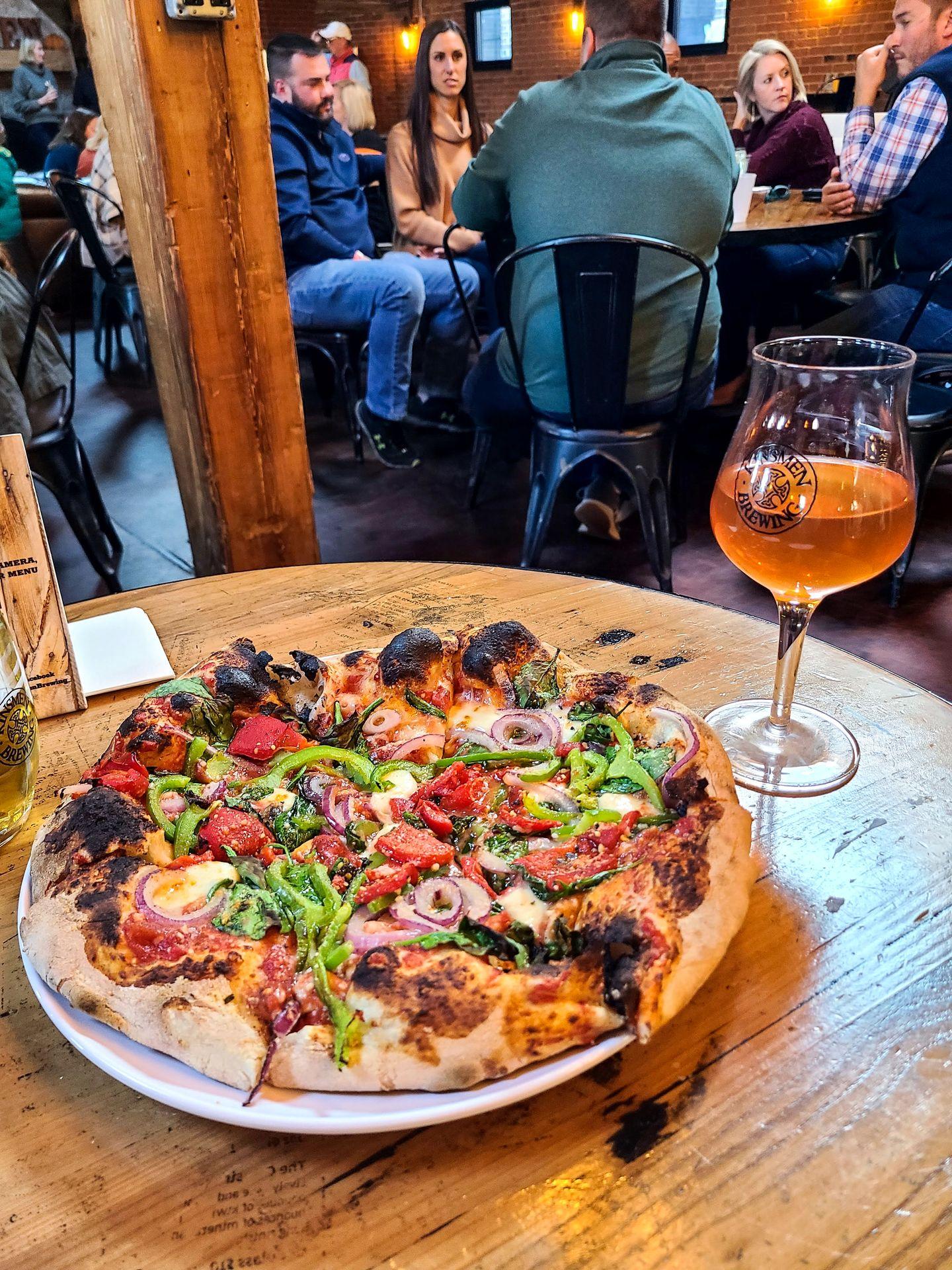 A pizza topped with veggies and a beer in a wine glass next to it on a table.