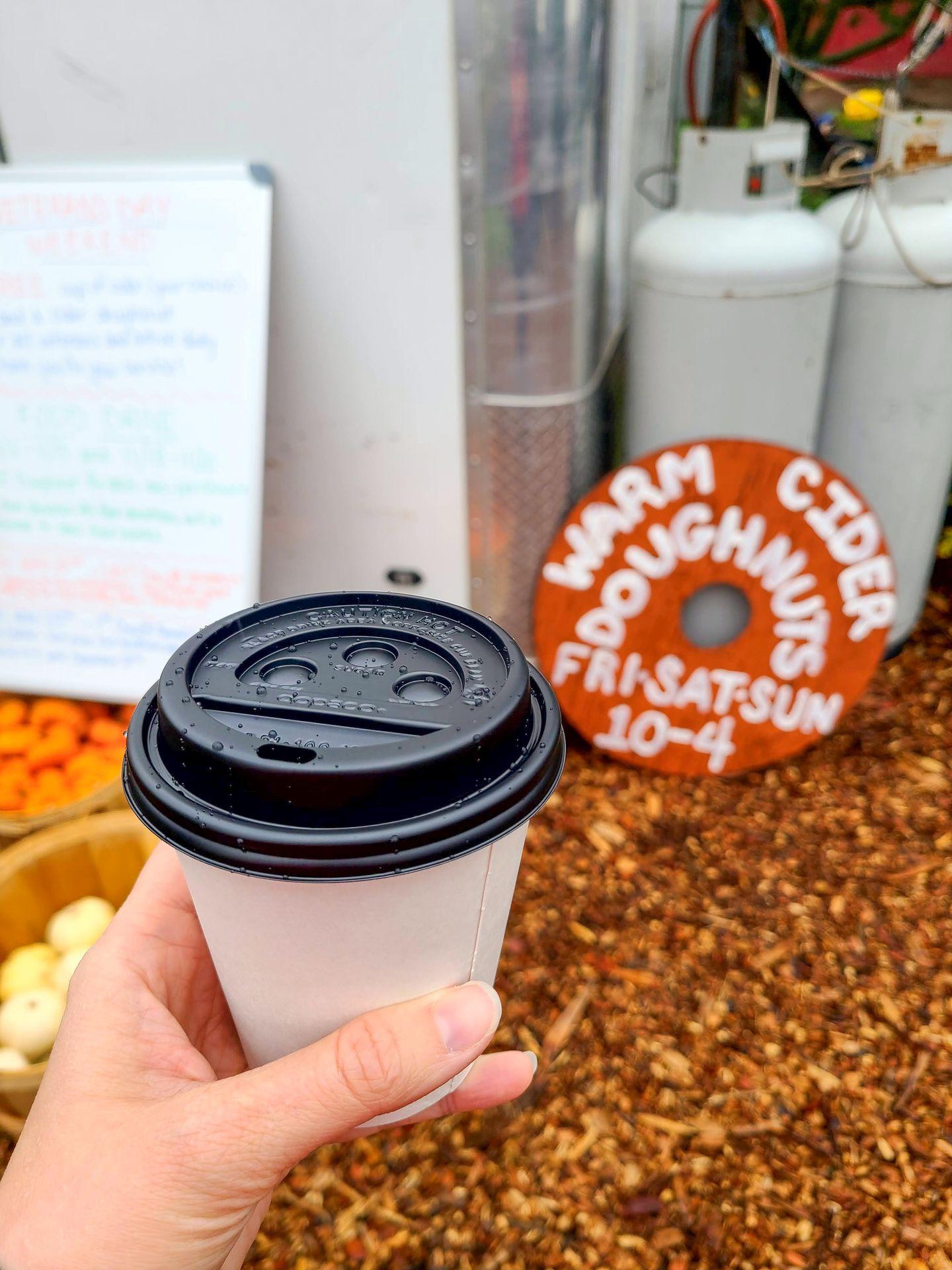 Holding a cup of hot apple cider. A sign in the background reads 'Warm cider doughnuts fri-sat-sun 10-4'